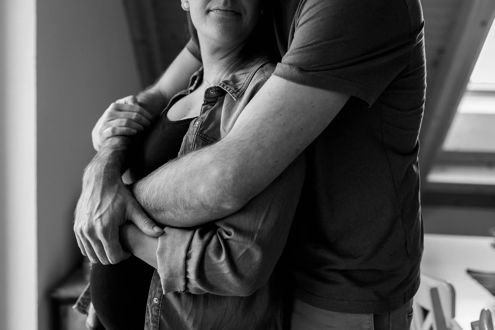 Nahaufnahme eines Mannes, der eine Frau umarmt, nur teilweise sichtbar, in einem Raum mit natürlichem Licht, schwarz-weiss
