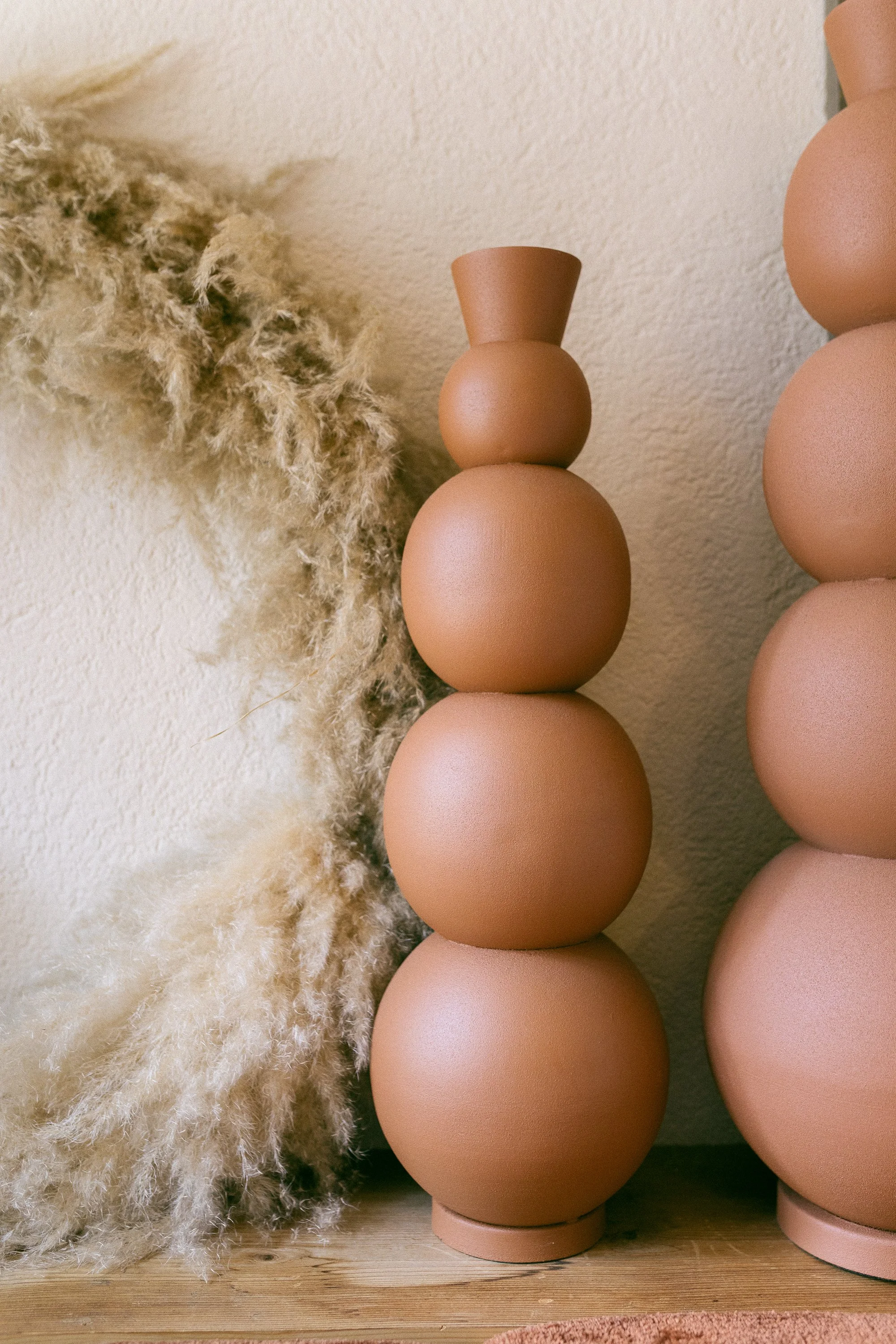 Braun-rote Keramikvasen neben einem Pampas-Kranz vor einer Holzoberfläche.