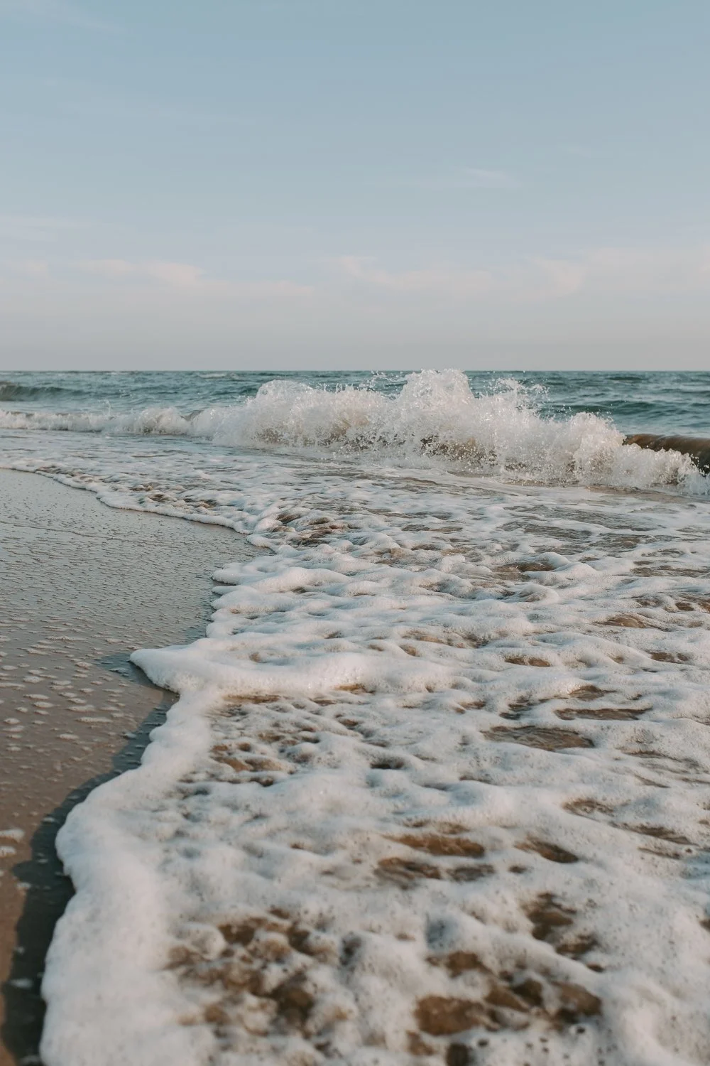 Aufnahme eines Meeresstrandes mit schäumendem Wellengang, Sand im Vordergrund, blauer Himmel im Hintergrund.