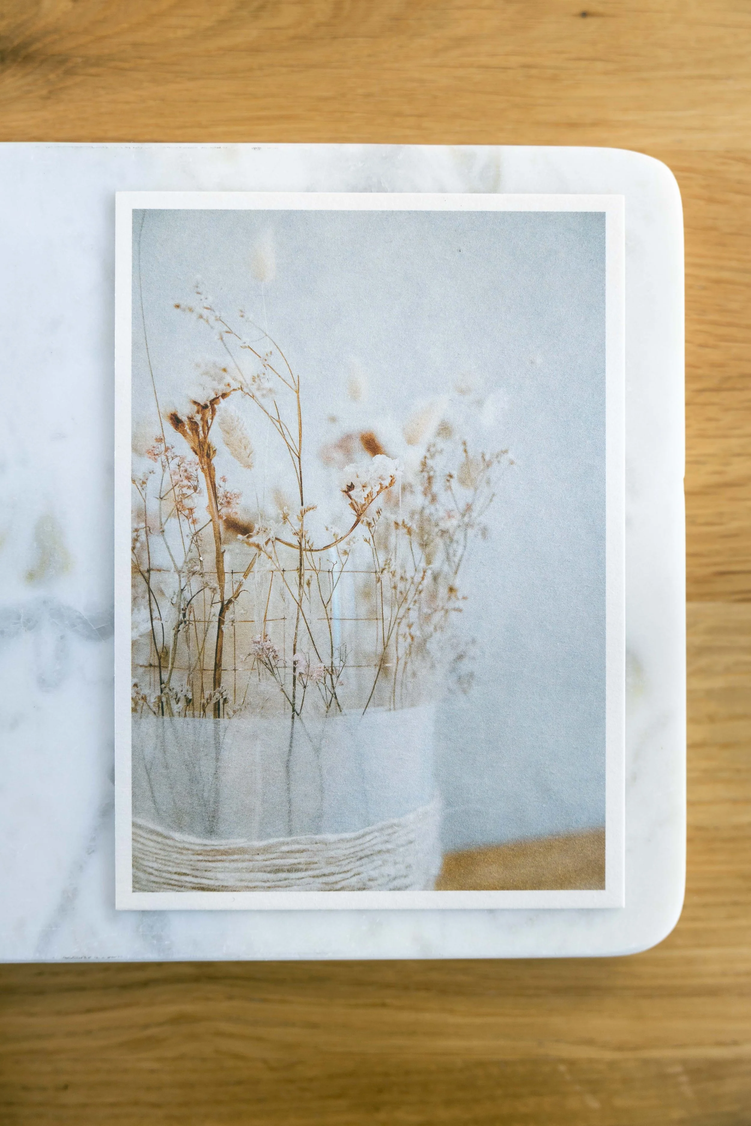 Fotografie einer Pflanzen-Postkarte mit trockenem Blumenstrauß in einer weissen Vase, auf einem Holztisch.