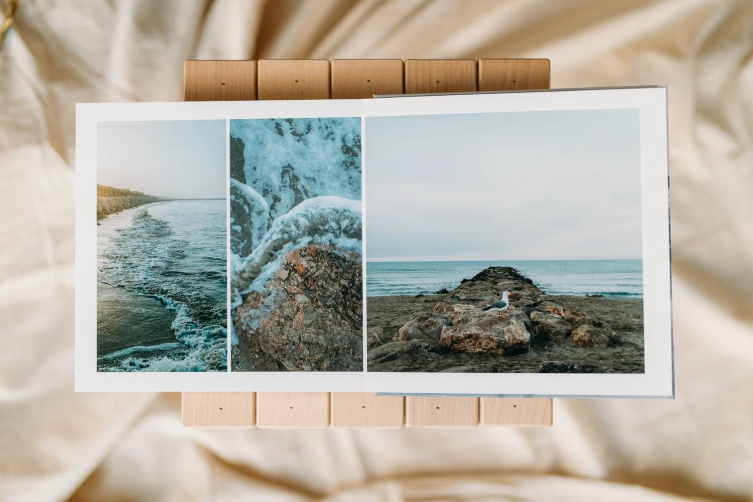 Foto eines geöffneten Fotoalbums mit Bildern des Meeres, Wellen und Felsen. Das Album liegt auf einem hellen Stoffhintergrund.