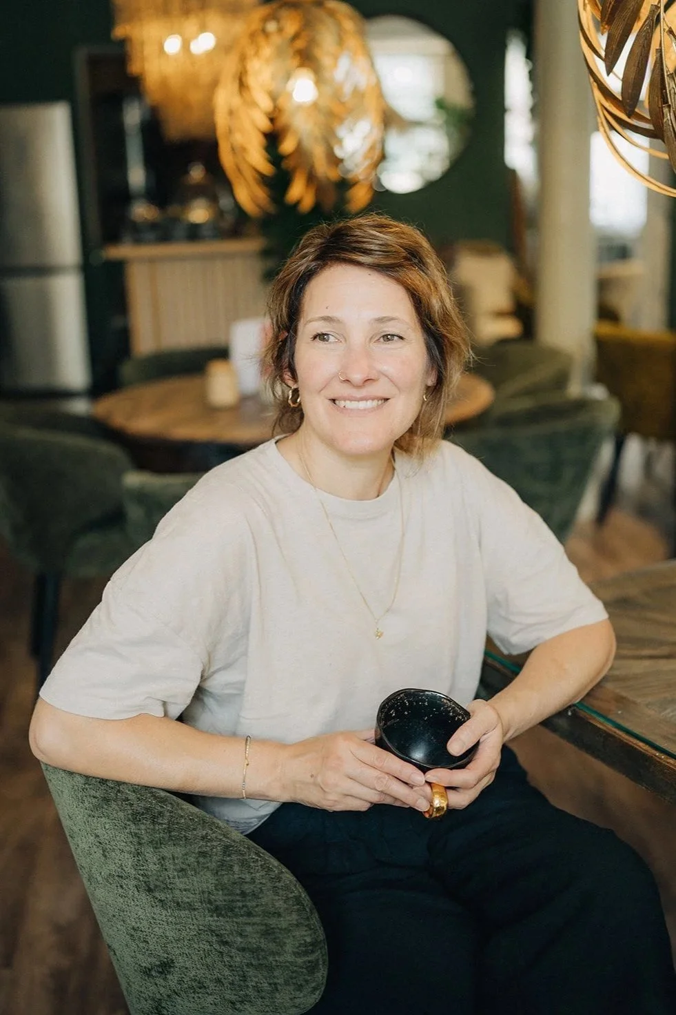 Eine lachende Frau mit kurzen braunen Haaren sitzt in einem Café, hält eine schwarze Tasse in den Händen, trägt eine beige T-Shirt, goldene Schmuckstücke und sitzt auf einem grünen Stuhl.