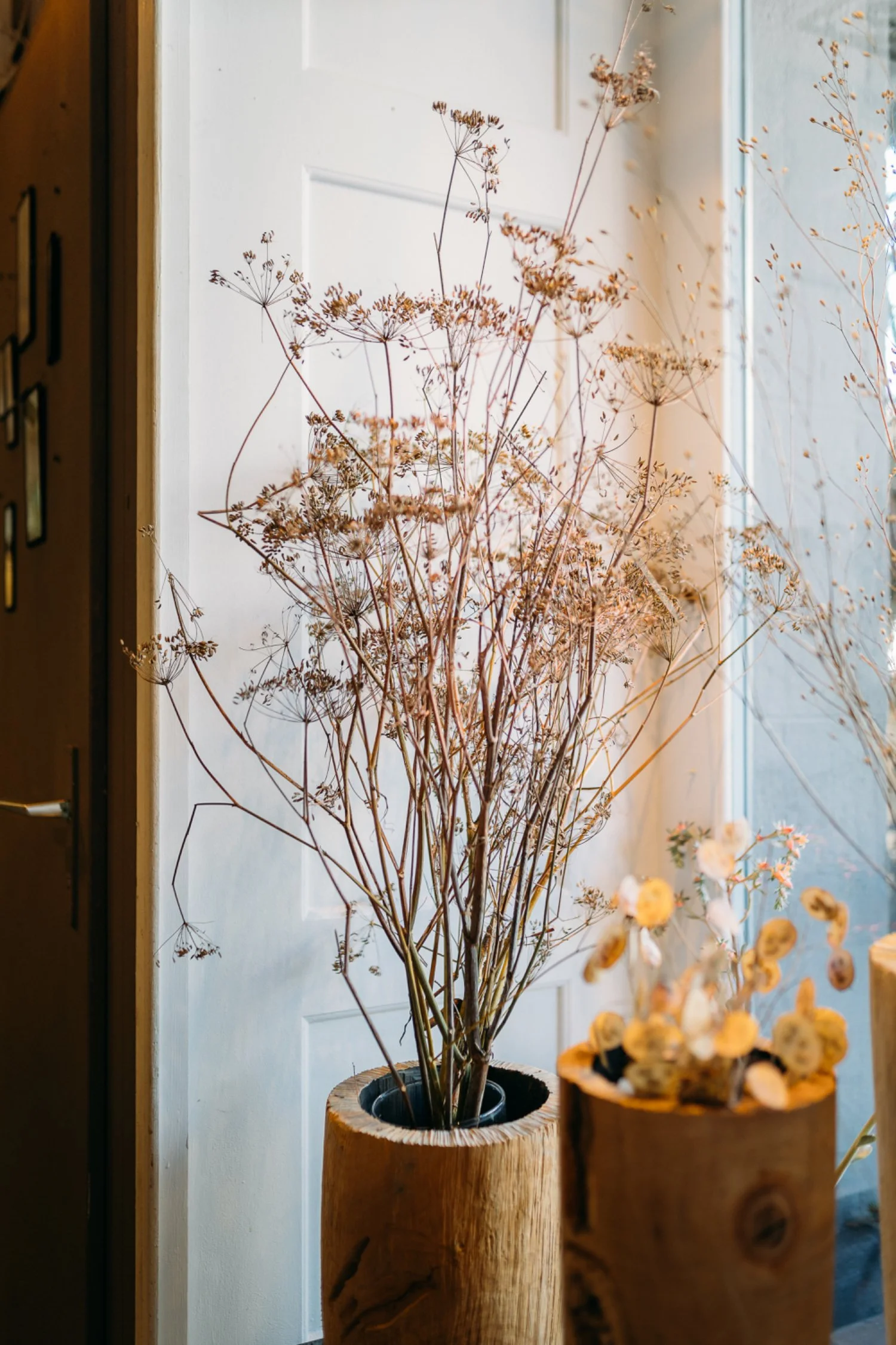 Tausende trockene Blätter und Zweige in einer grossen Holzvase neben einem anderen Holzbehälter mit kleineren trockenen Pflanzenteilen, vor einem Fenster.