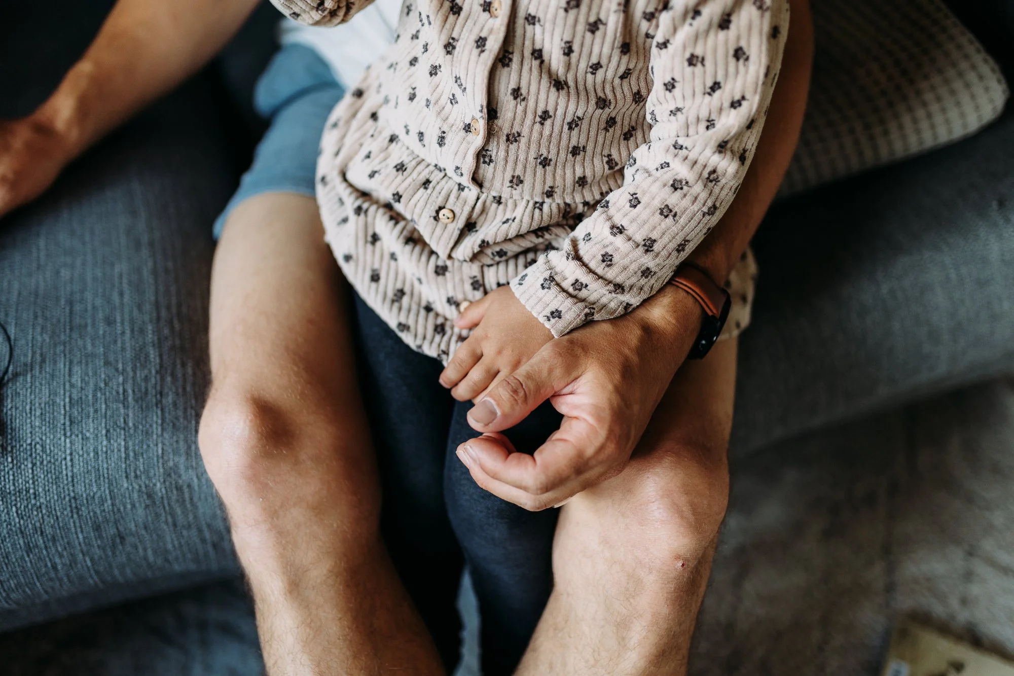Ein Erwachsener hält ein kleines Kind auf dem Schoß, während sie auf einem grauen Sofakissen sitzen. Das Kind trägt ein beige Hemd mit kleinen schwarzen Blumenmustern.