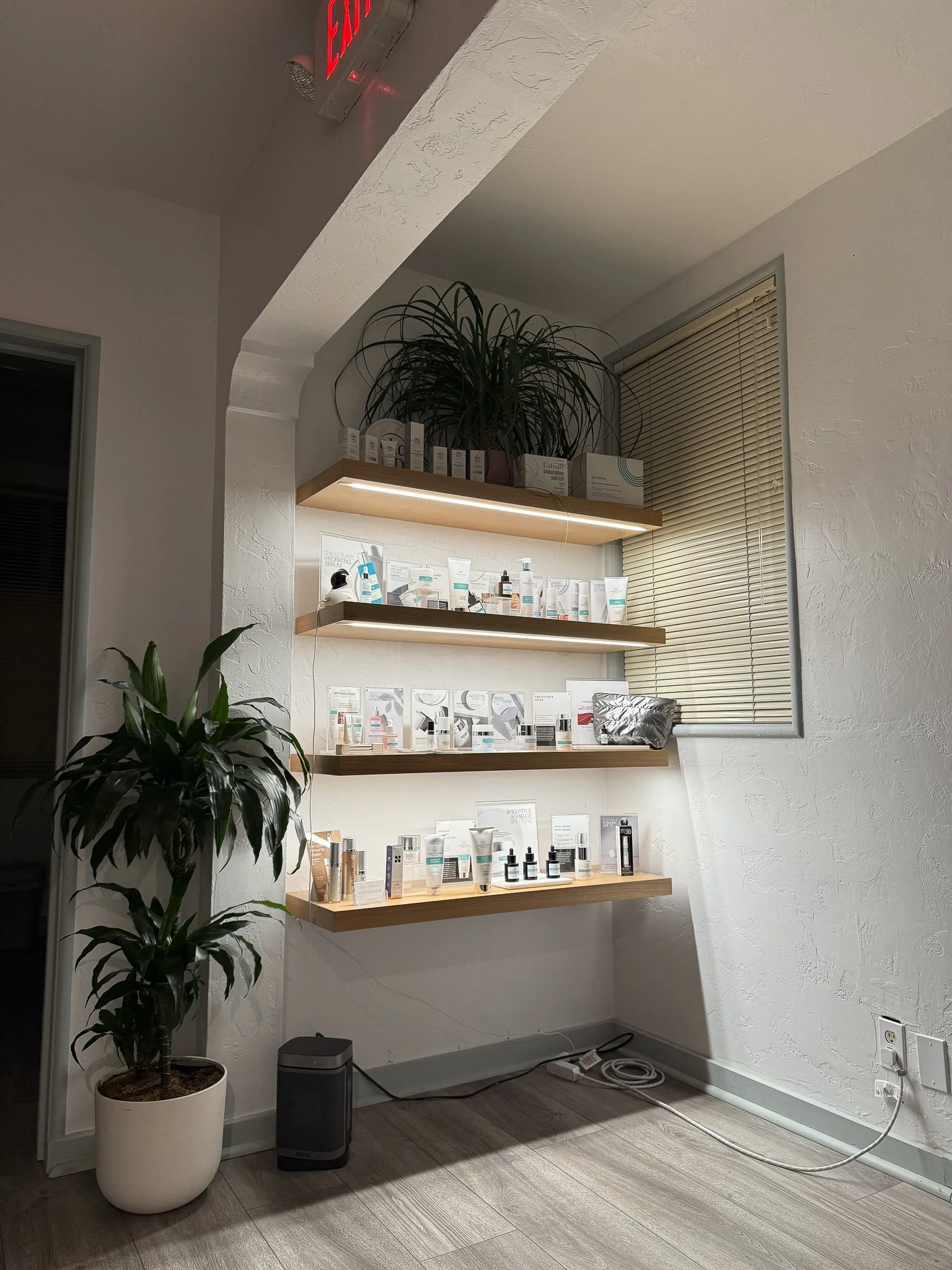 Interior of a skincare retail space with three wooden shelves with built in LED strips lighting skincare products, a large potted plant on the left, a window with closed blinds on the right, and a vent with an exit sign on the ceiling