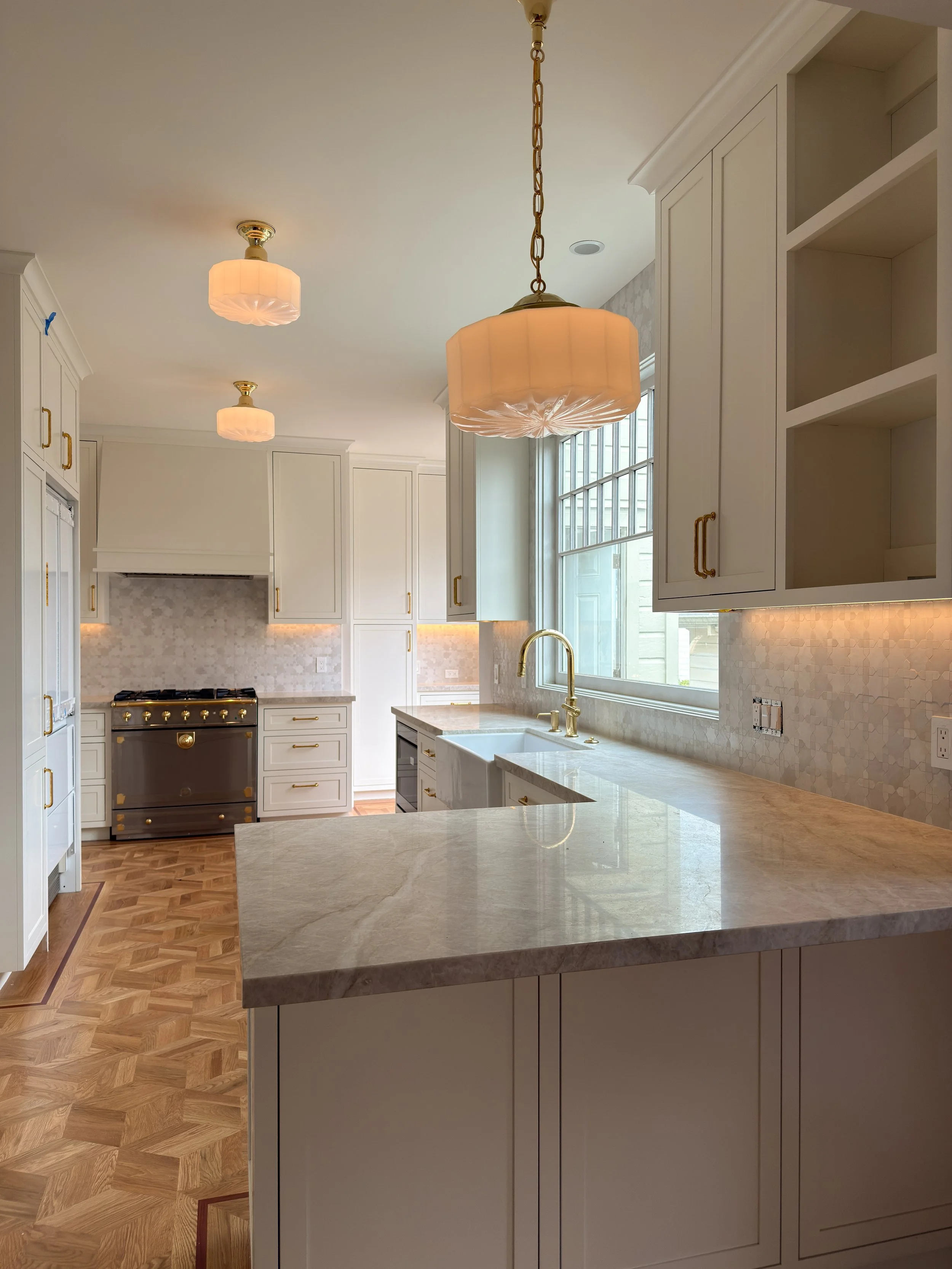 Modern kitchen with white cabinets, marble countertops, brass hardware, large window, and pendant lighting. There's beautiful undercabinet lighting, as well as classic surface mounted lights
