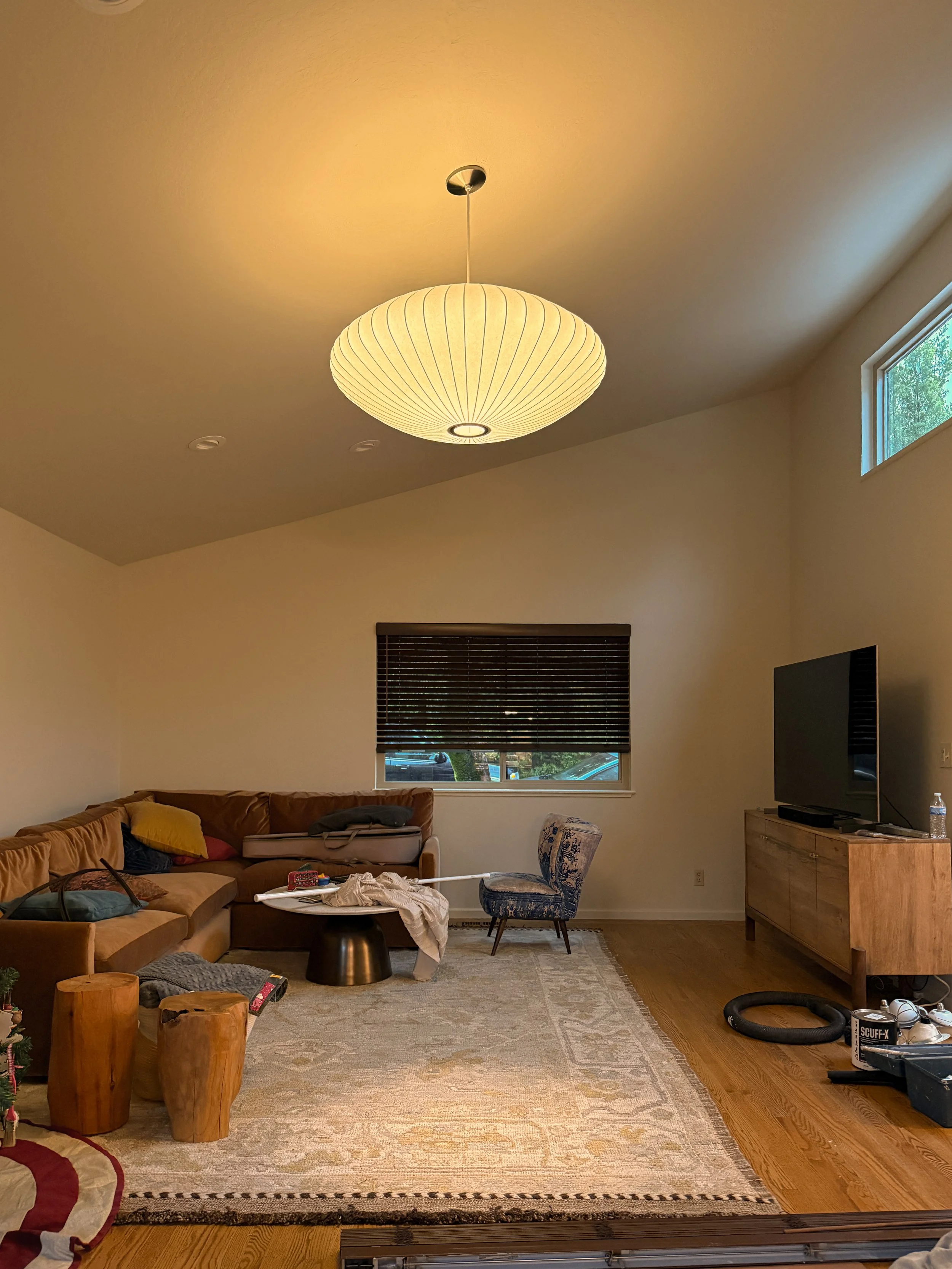 Living room with beige walls, a brown sectional sofa with colorful pillows, a patterned area rug, a window with blinds, a wooden cabinet with a TV on top, and various scattered items including a vacuum hose, a box, and miscellaneous objects.