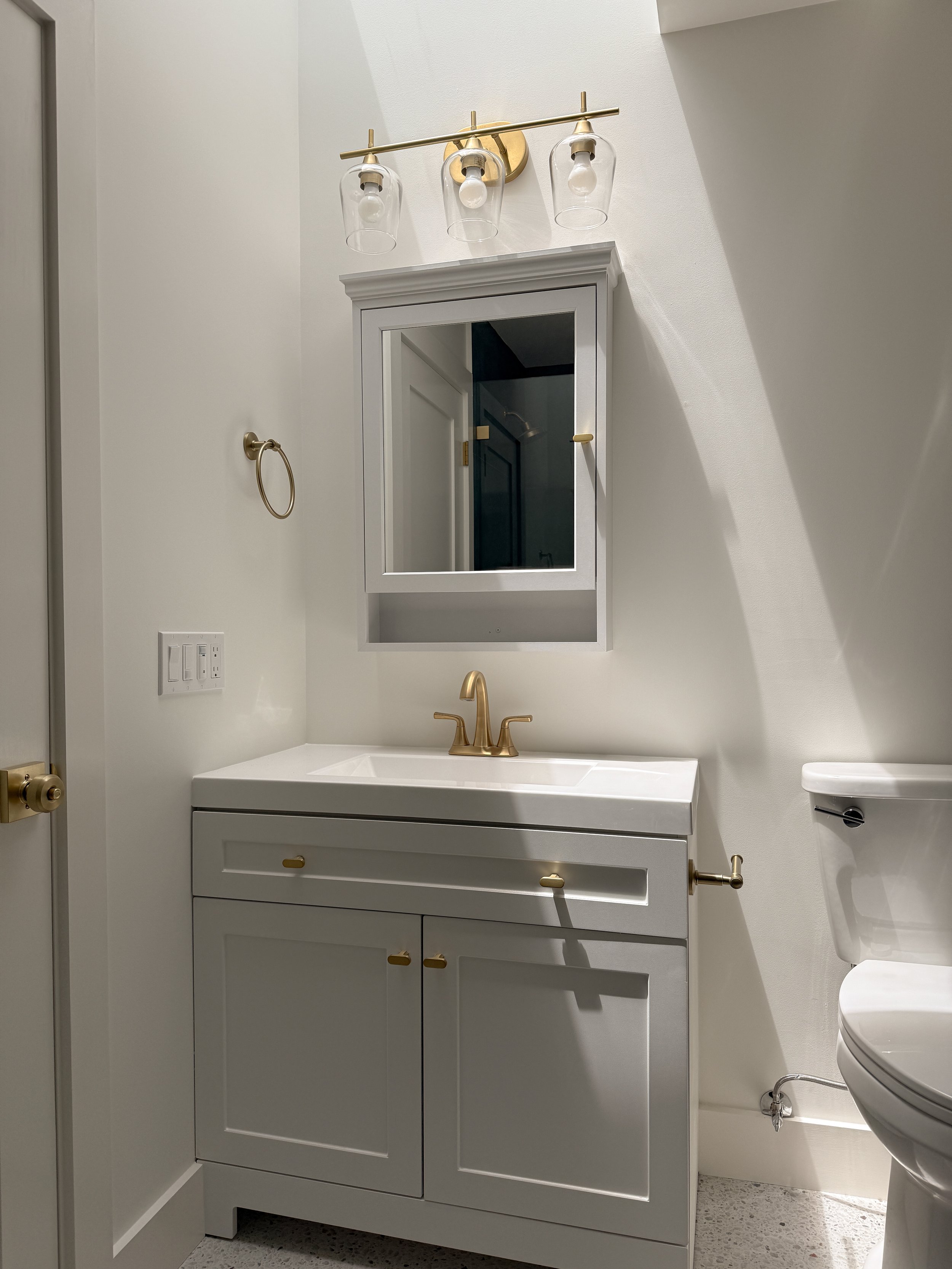 A small modern bathroom with a white vanity cabinet, gold fixtures, and a large mirror. There is a white sink with a gold faucet, a wall-mounted light fixture with three glass shades, a towel ring, and a partial view of a toilet. The bathroom has a light-colored floor and minimal decor.