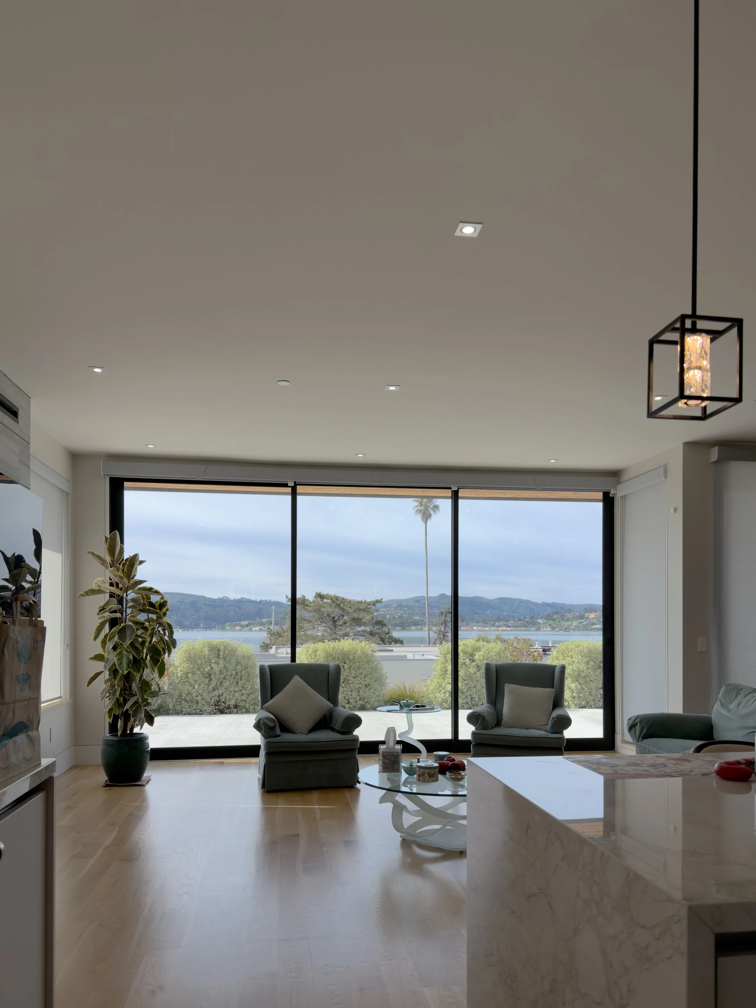 Living room with large glass doors overlooking a lake and mountains, two armchairs with pillows, side table, indoor plants, and modern lighting fixtures.