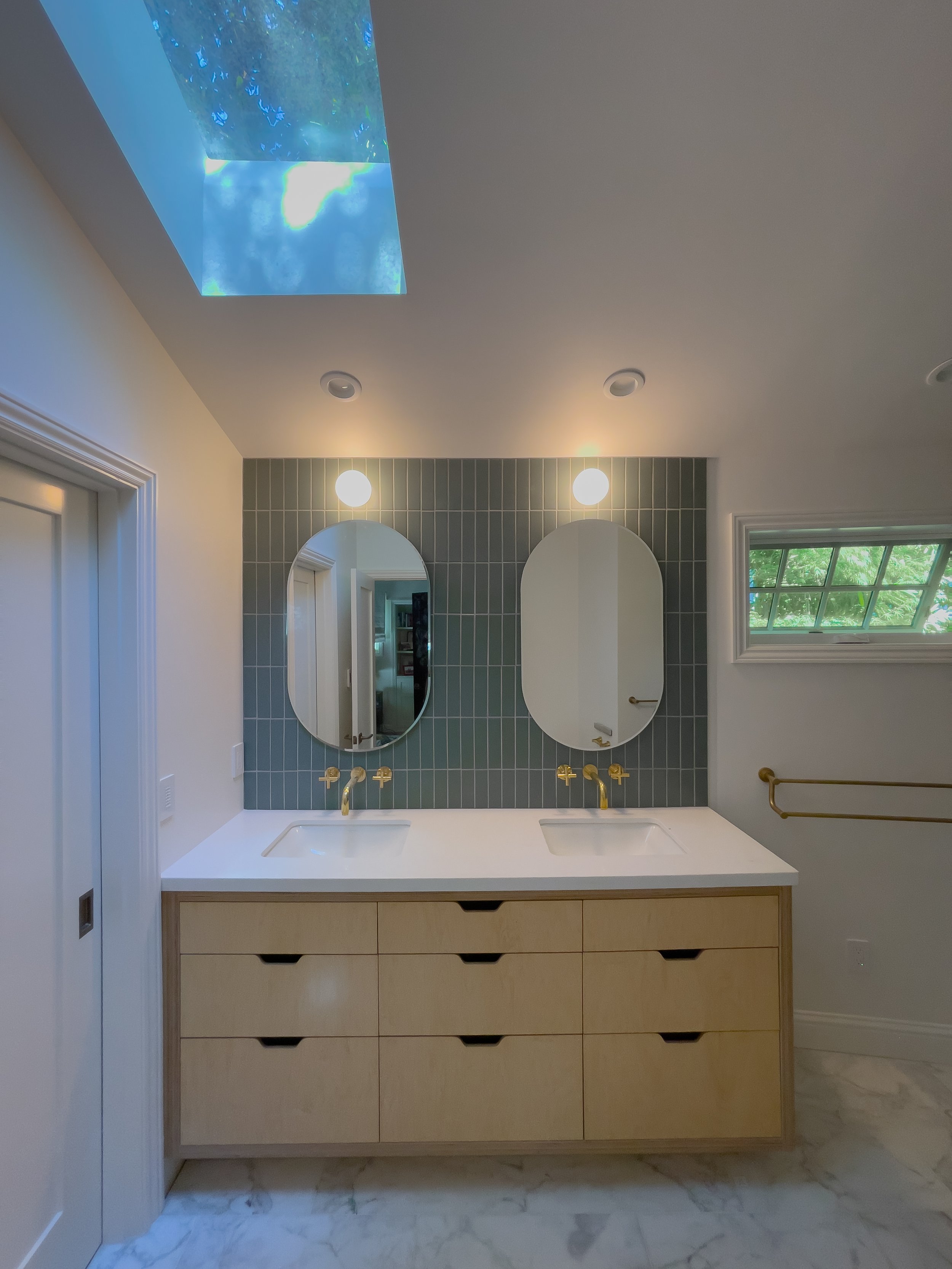 Modern bathroom vanity with double sinks, two oval mirrors, white countertop, gold fixtures, light gray tile wall, a window with greenery outside, and a skylight with blue sky reflection.