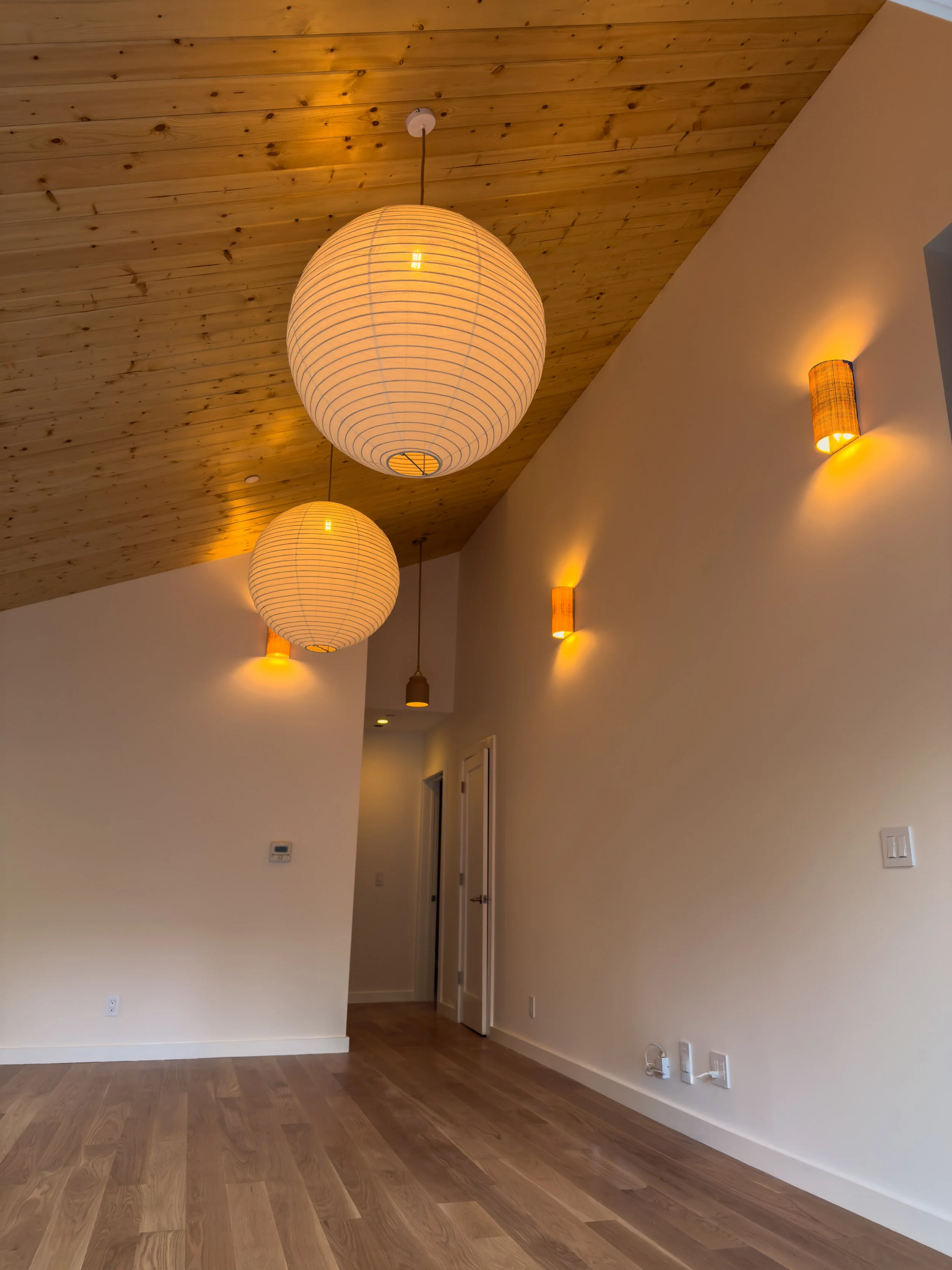 Image of a modern interior room with a wood-paneled sloped ceiling, white walls, brown wood flooring, and various wall-mounted sconce light fixtures providing warm lighting, as well as large round pendants 