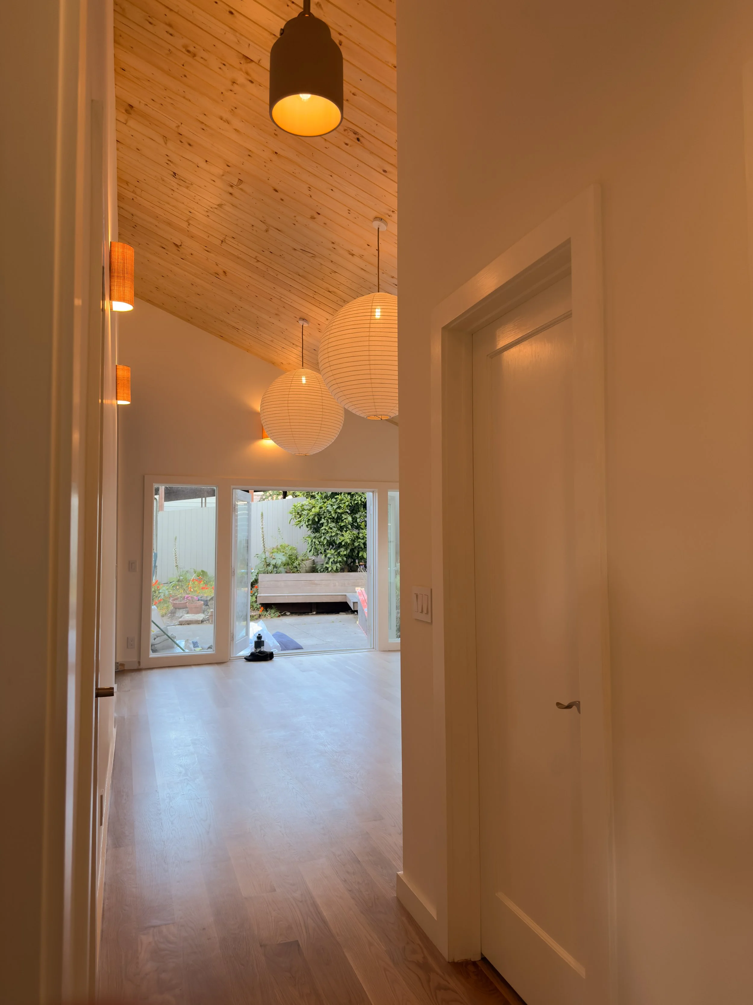 Interior view of a room with vaulted wooden ceilings, hanging pendant lights, sconces installed on wall, and sliding glass doors leading outside to a garden.