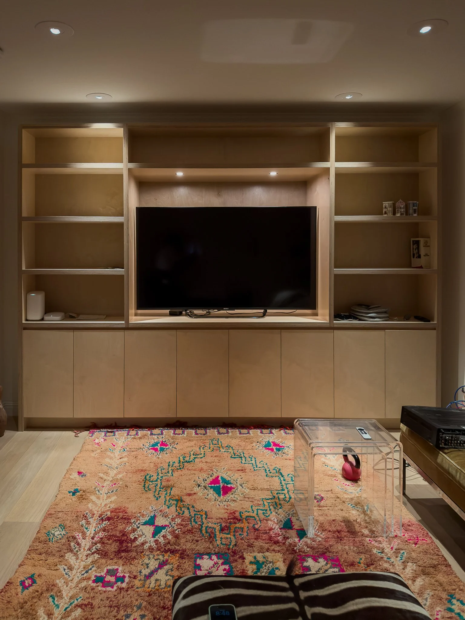 A room with a large built-in wooden entertainment center with open shelves, a flat-screen TV that hides ethernet connections behind it, a colorful area rug, and a clear acrylic side table. The ceiling has recessed lights that were upgraded to pinhole