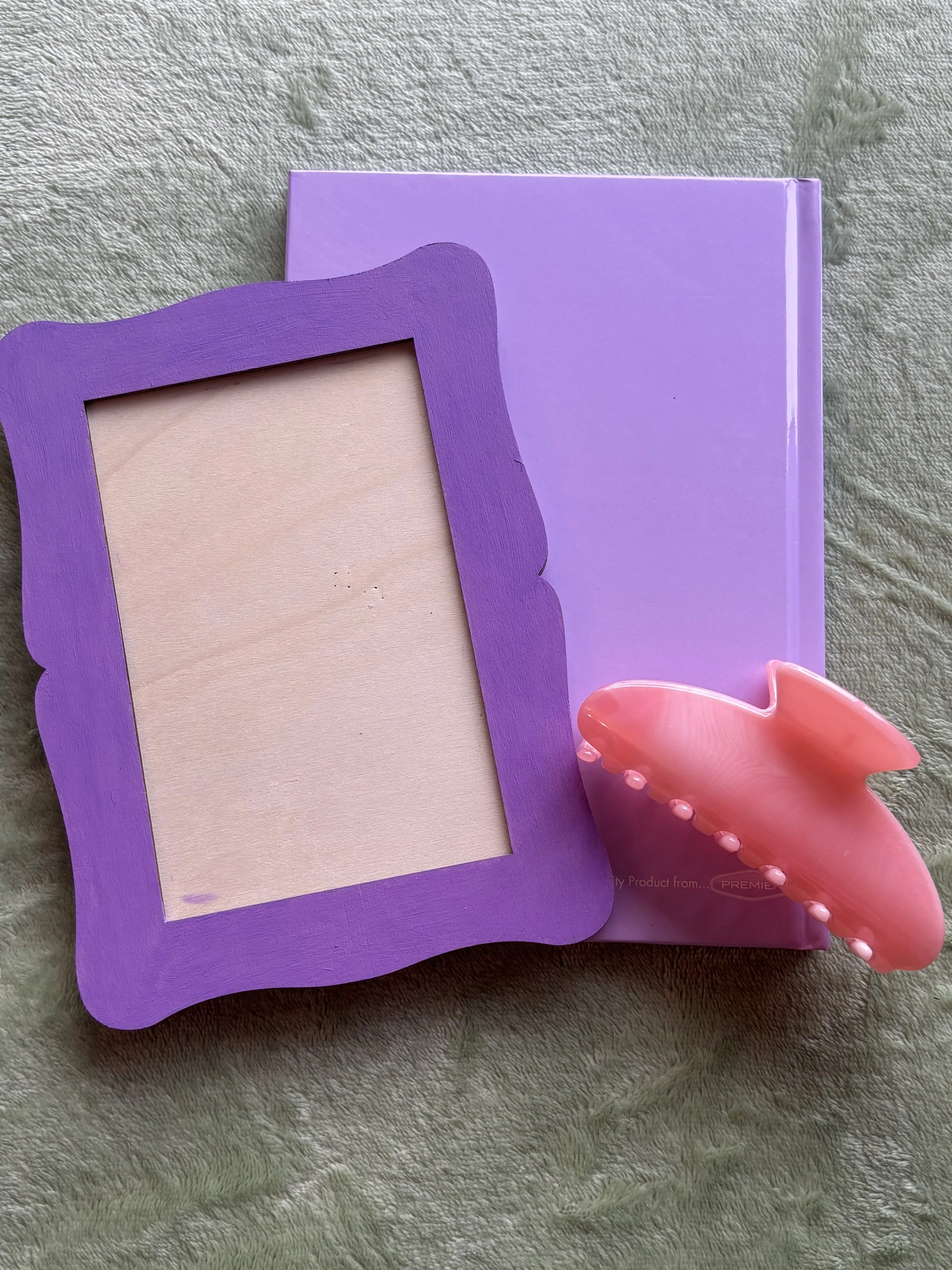 A purple picture frame, a purple card with rounded edges, and a pink dolphin-shaped hairbrush with beads along the side are placed on a soft, textured gray surface.