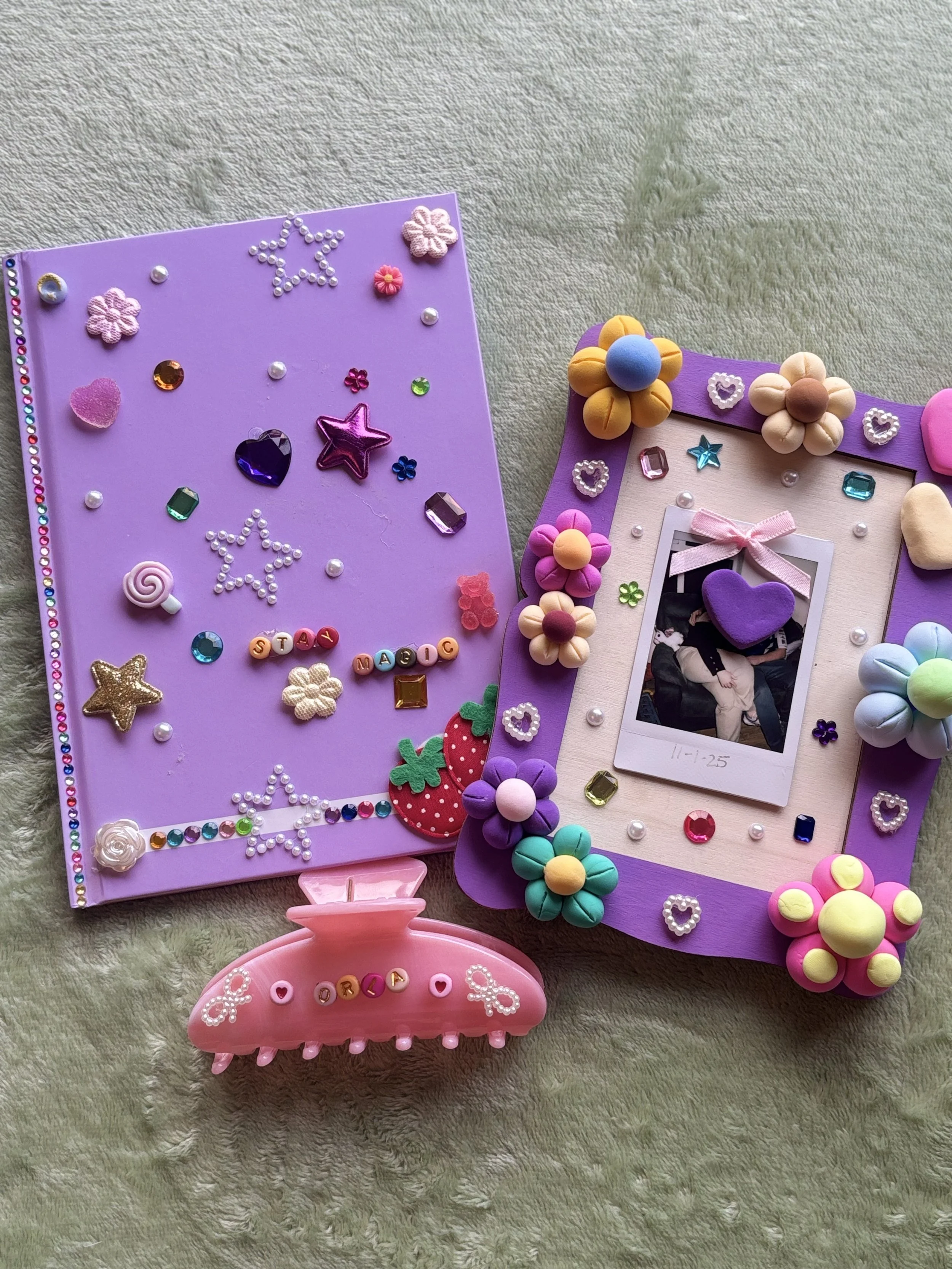 Decorative photo frame with flowers, hearts, and beads, and a small picture inside. A pink hair clip with the word 'DORA' in colored beads is below in the picture. The background is a beige textured surface.