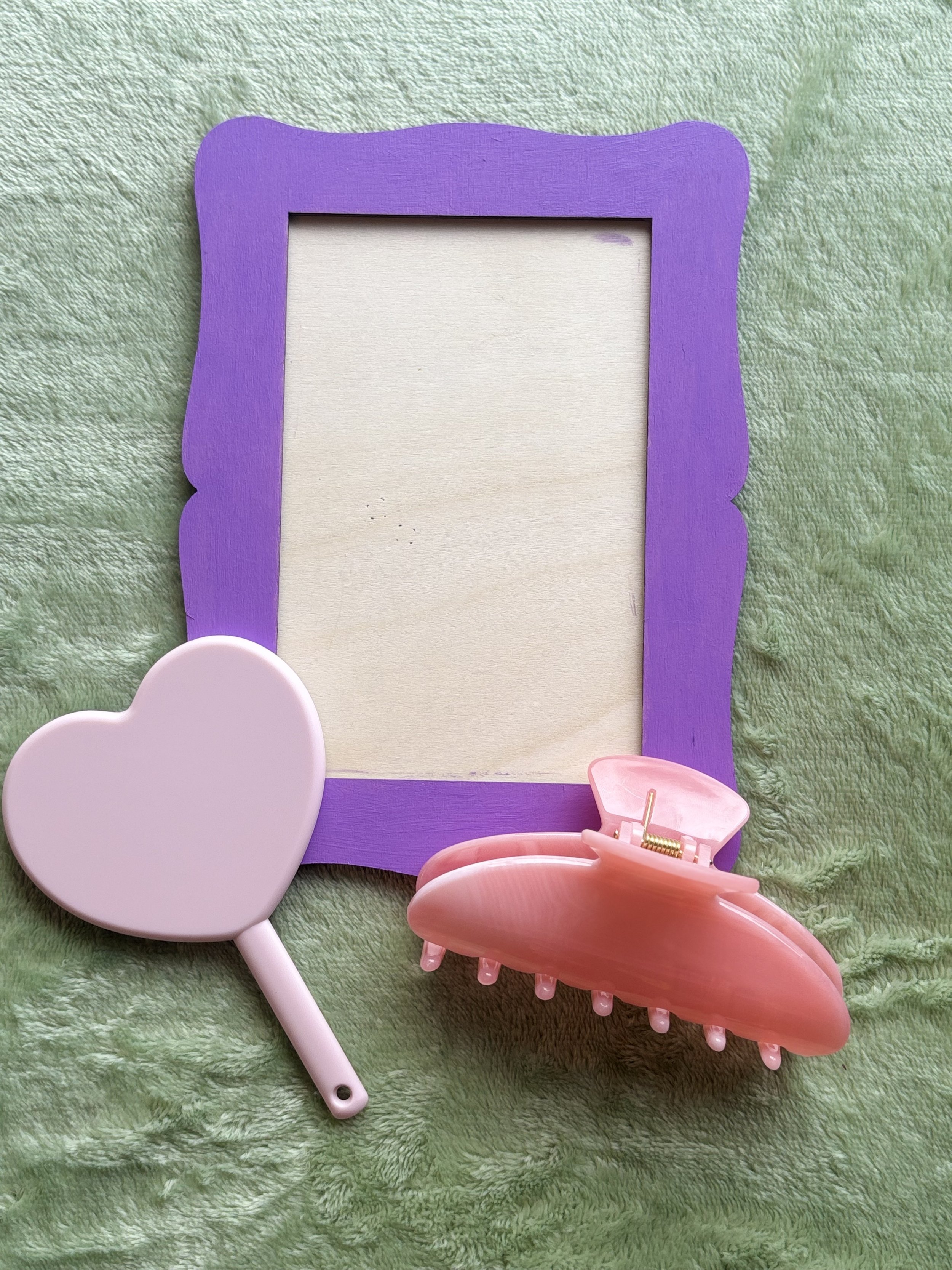 A collection of pastel-colored toy accessories including a purple frame, a pink sun hat, a pink hair clip, and a pink mirror, all arranged on a green textured fabric background.
