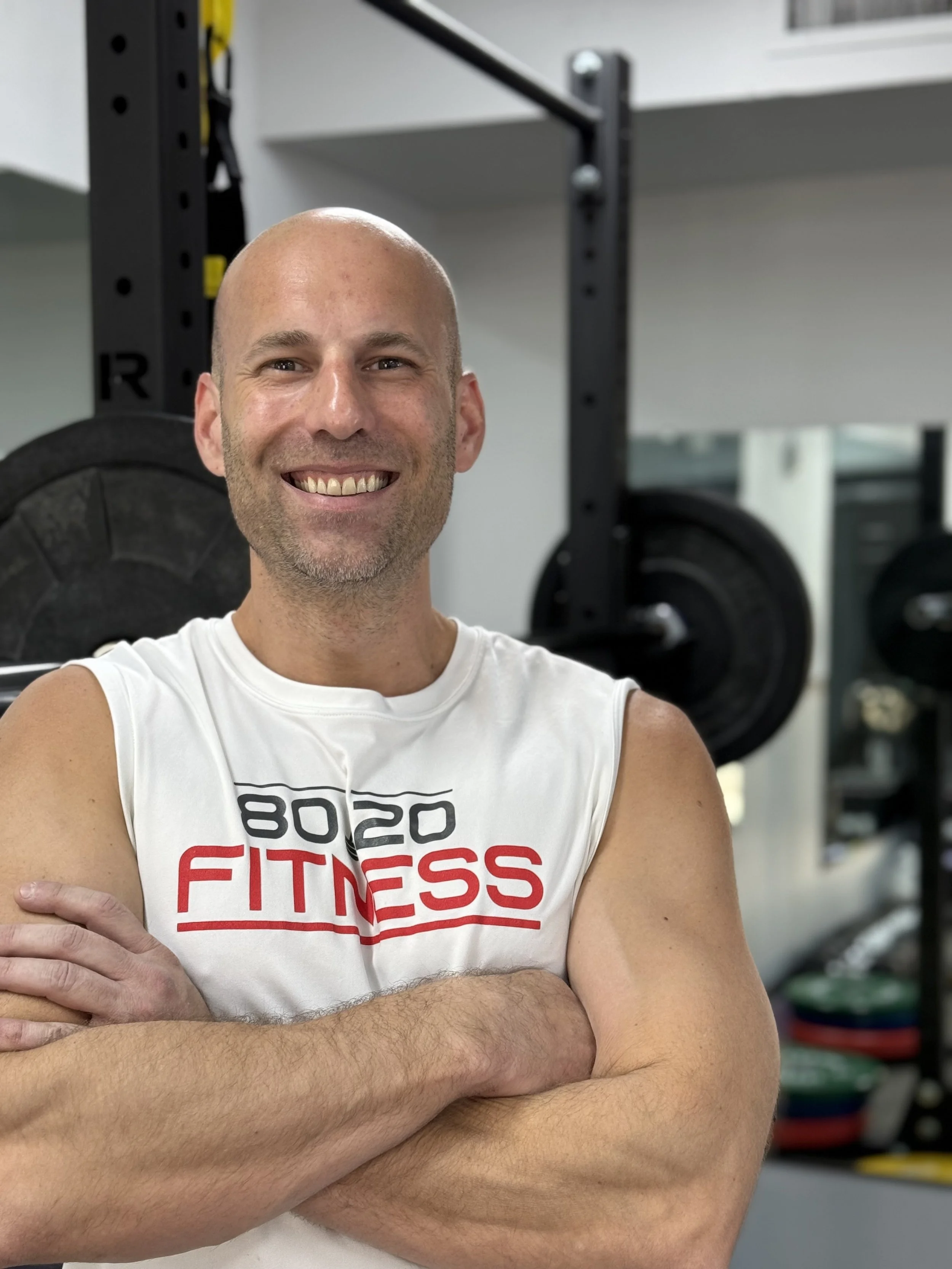 A smiling man with a shaved head and beard in a gym, wearing a white sleeveless shirt with the text '8020 FITNESS', crossed arms, gym equipment including weights in the background.
