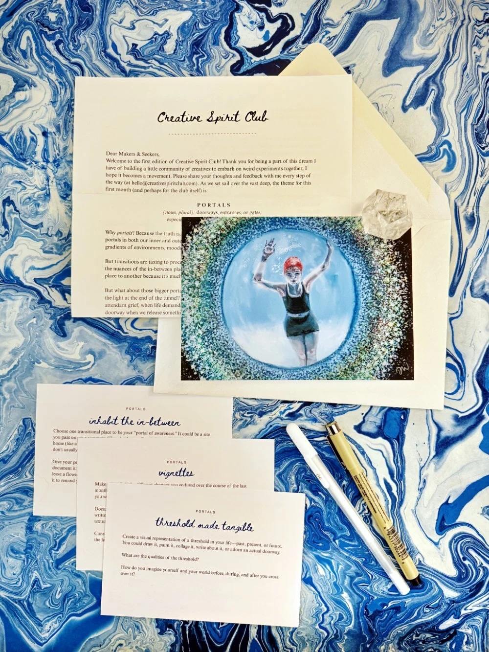 Collection of printed papers and a colorful artwork, placed on a marbled blue and white surface, with two pens and a crystal on the papers.