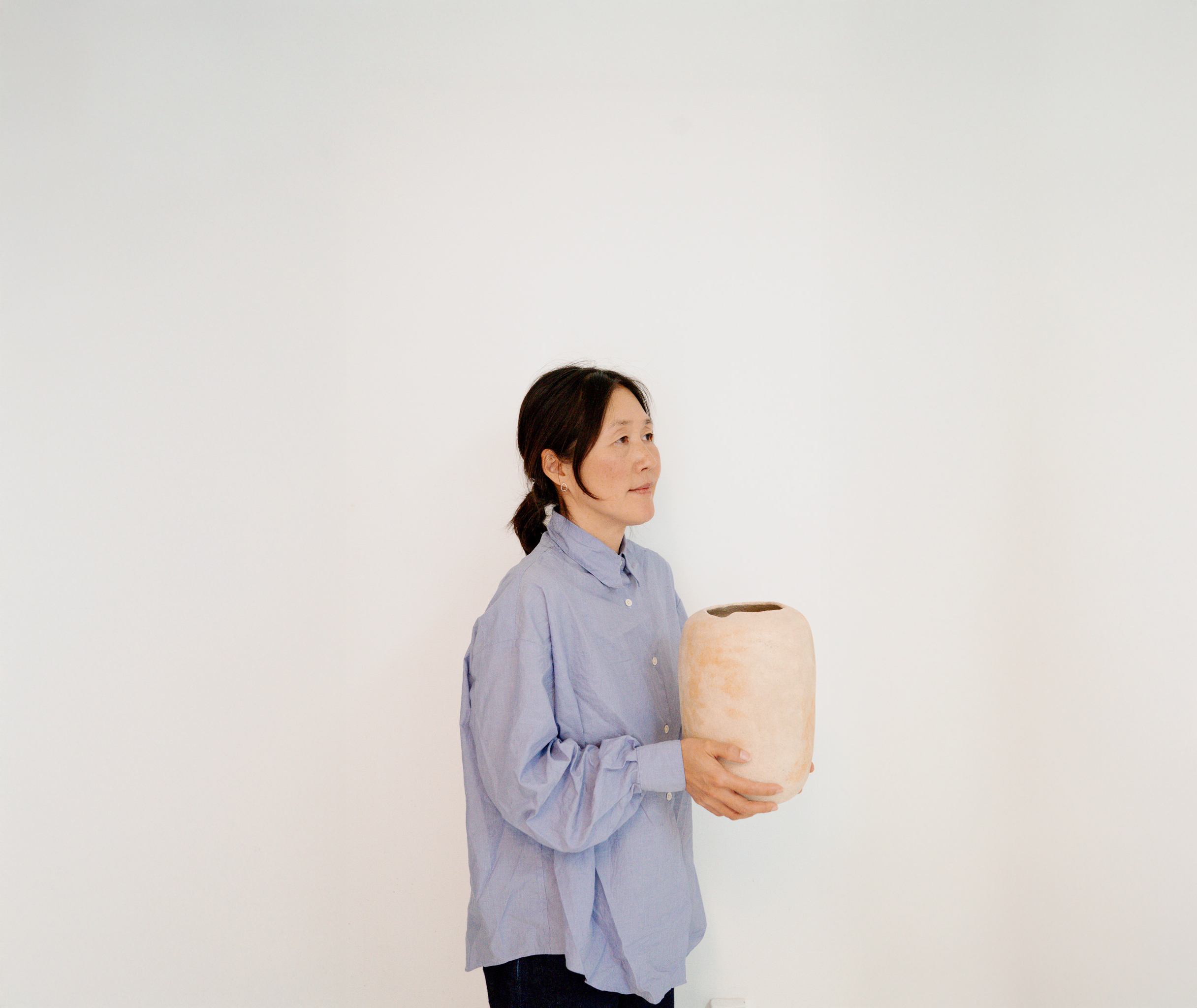 Woman in blue shirt holding a beige vase against a white background.