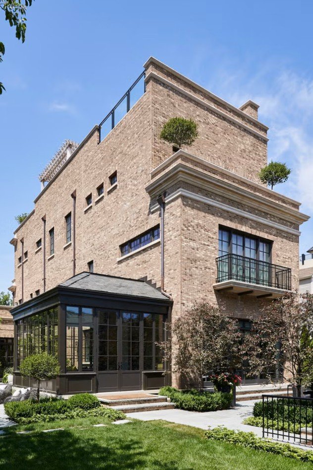 A modern multi-story brick house with a glass sunroom, balcony, and top terrace with small trees, set against a blue sky.