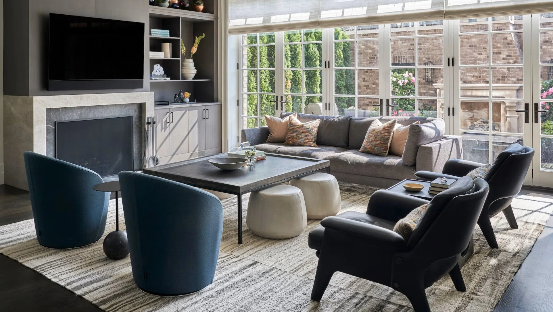 Living room with large windows, gray sofa with pillows, black armchairs, a rectangular coffee table, and a built-in fireplace with a TV above. Sunlight streams through the windows, illuminating the space.