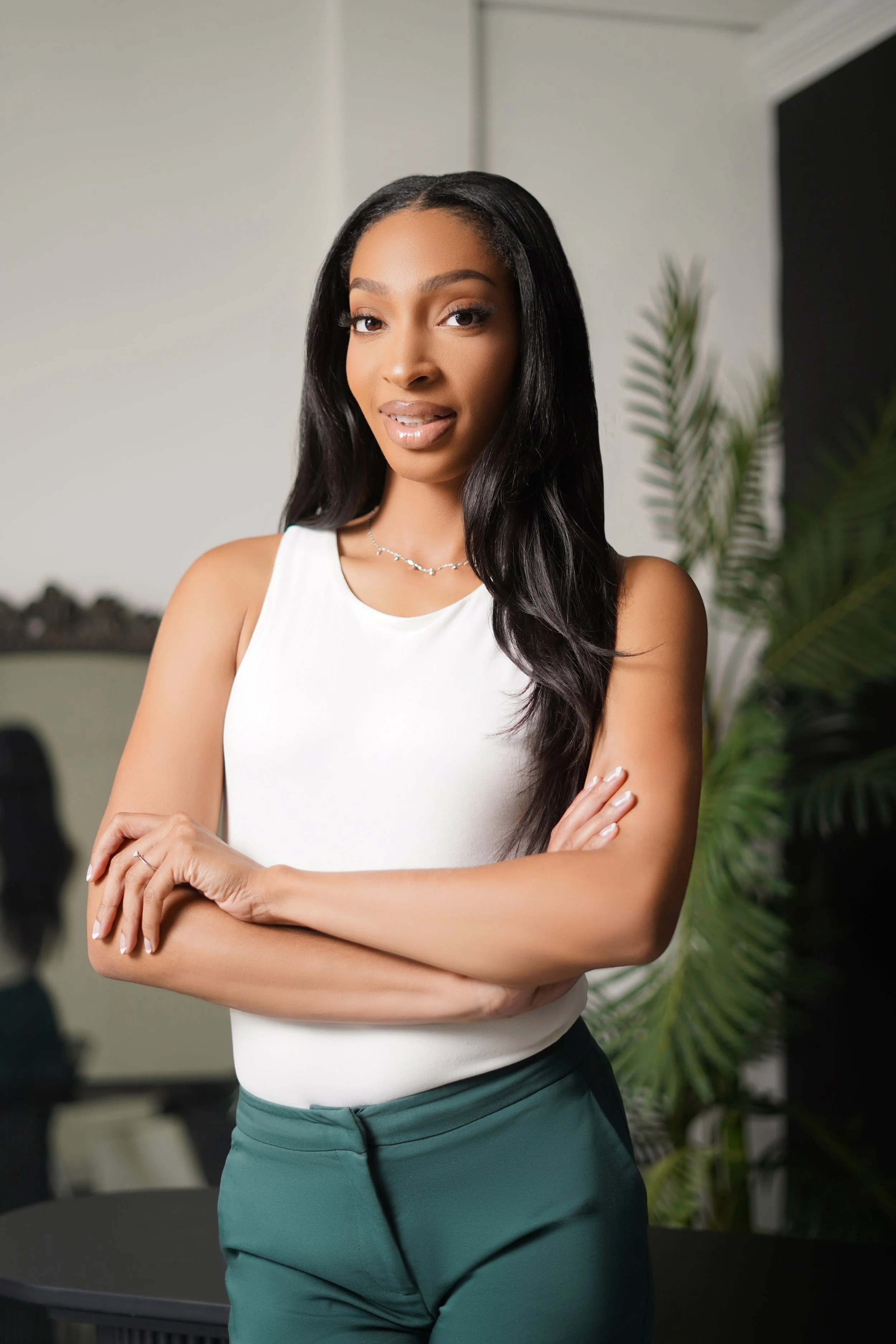 A confident young woman with long black hair, wearing a white sleeveless top and green pants, standing with her arms crossed in an indoor office setting with a plant and a blurred background.