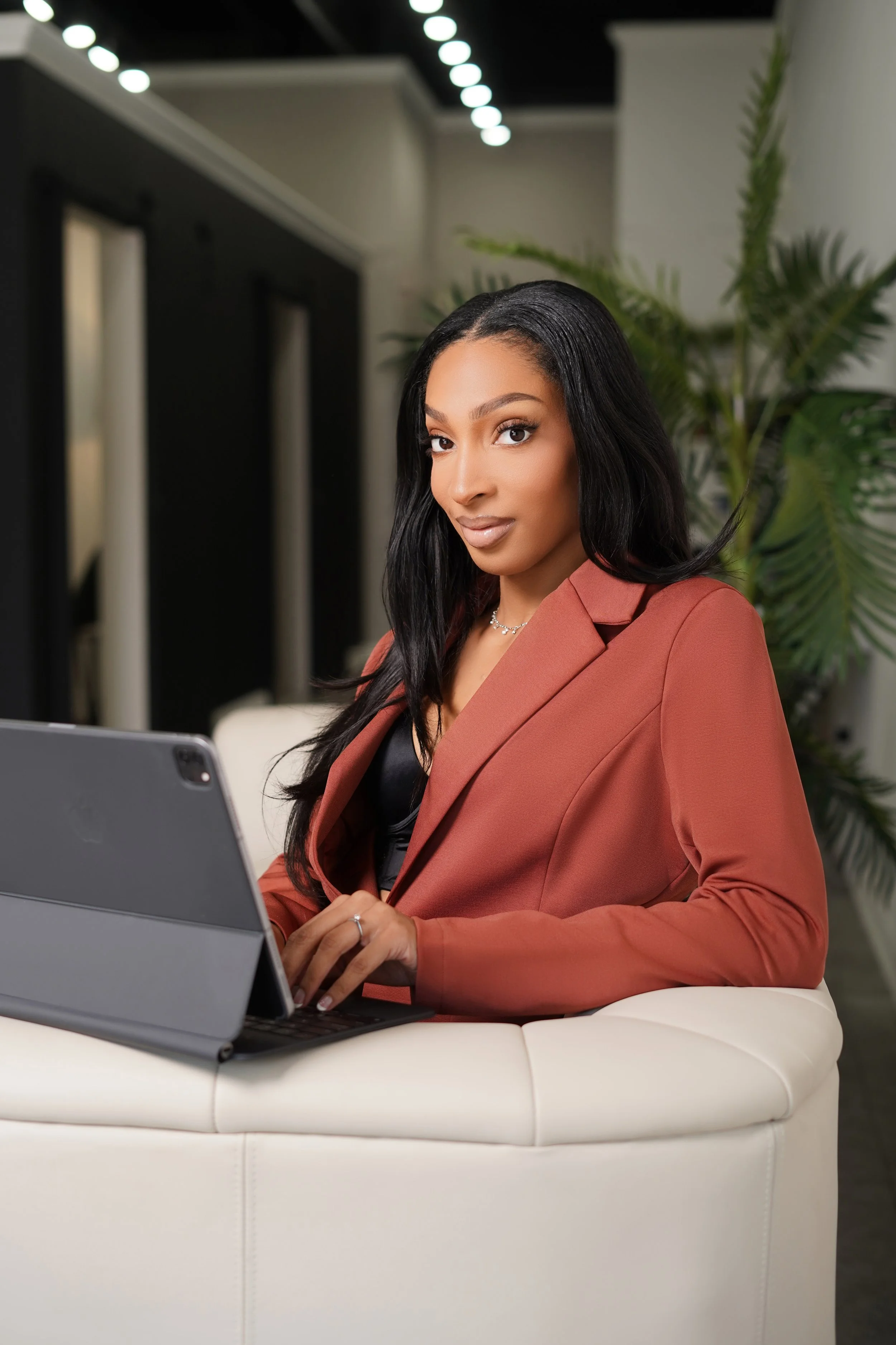 A woman sitting on a white couch using a tablet in an indoor setting with plants and modern decor.