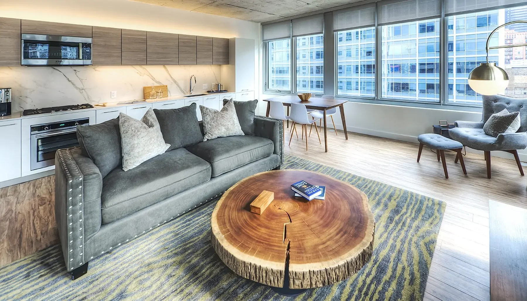 Modern living room with a gray sofa, a large wooden slab coffee table, a gray armchair with a matching footstool, a dining table with four white chairs, large windows with city views, and a kitchen area with white cabinets and a marble backsplash.