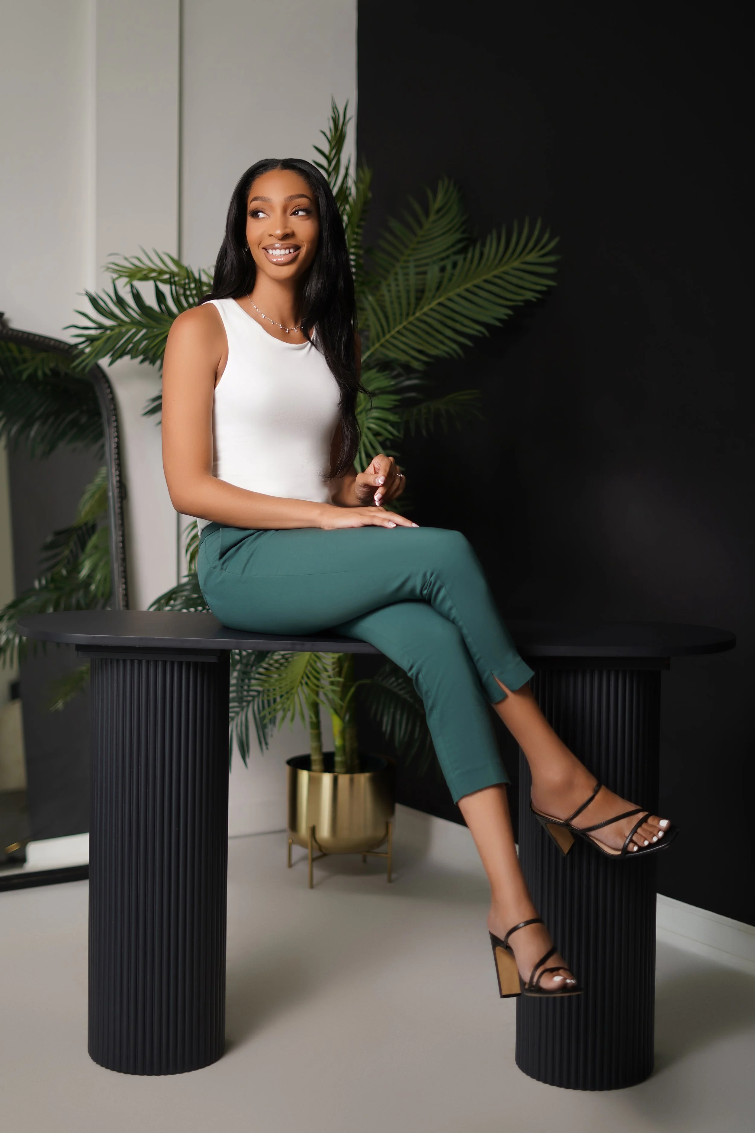 A woman in a white top and teal pants sitting on a black table with large black-striped legs, next to a potted plant with large green leaves in a gold pot, against a black and white wall.