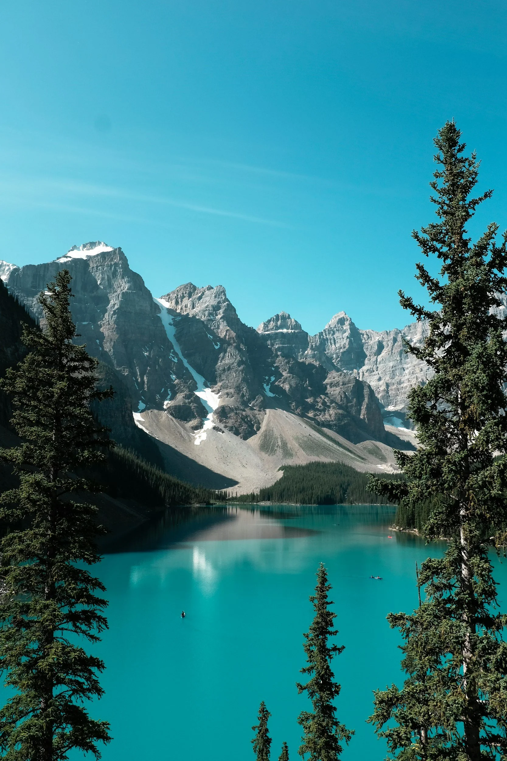 Scenic view of a turquoise lake surrounded by pine trees, with rugged snow-capped mountains in the background and a clear blue sky.