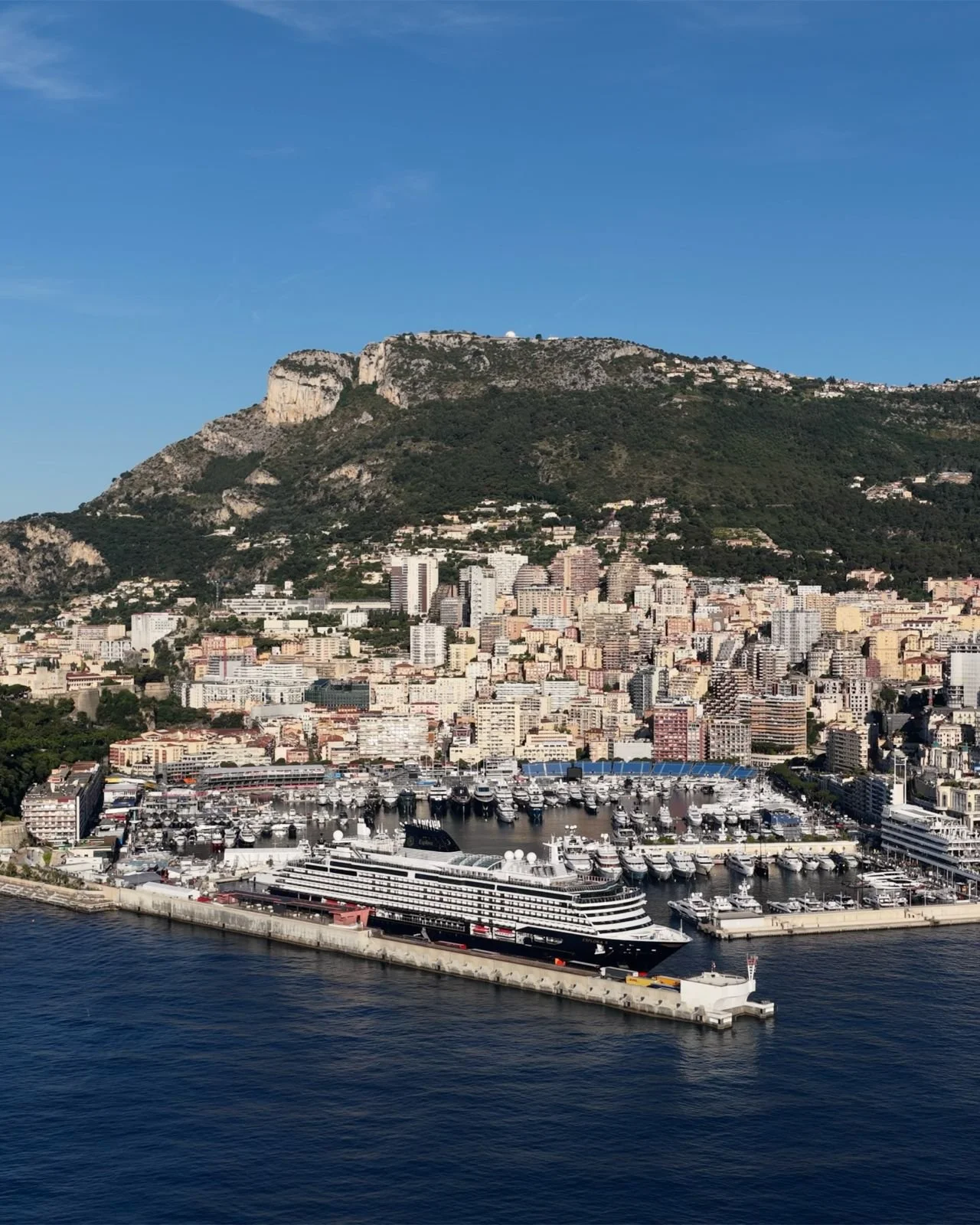 EXPLORA I docked in Monaco's Port Hercule, with the pastel buildings and mountains of Monte Carlo rising behind the harbor