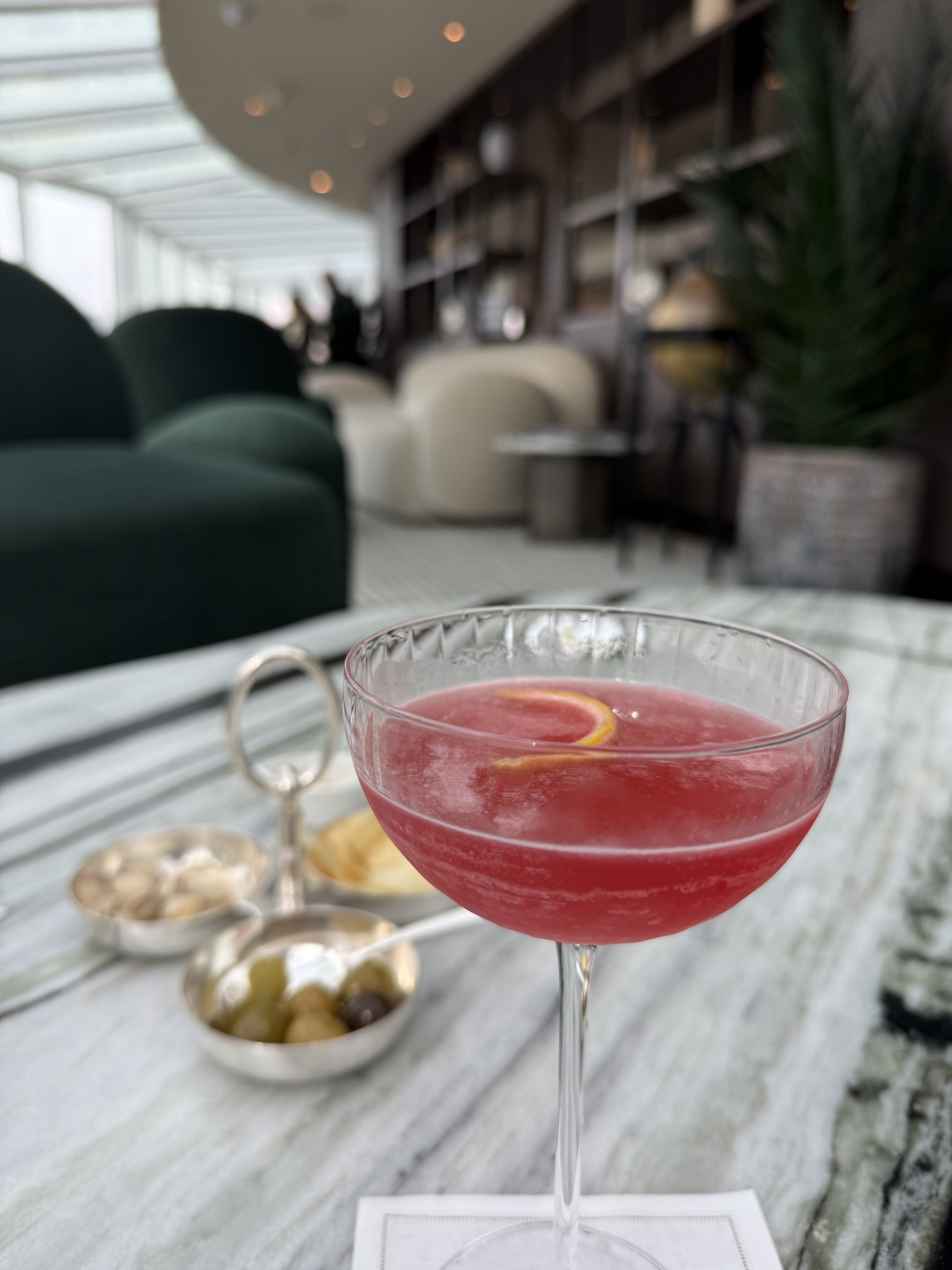 A blush-pink cocktail with a citrus twist and a dish of olives, served in a stylish lounge aboard EXPLORA I
