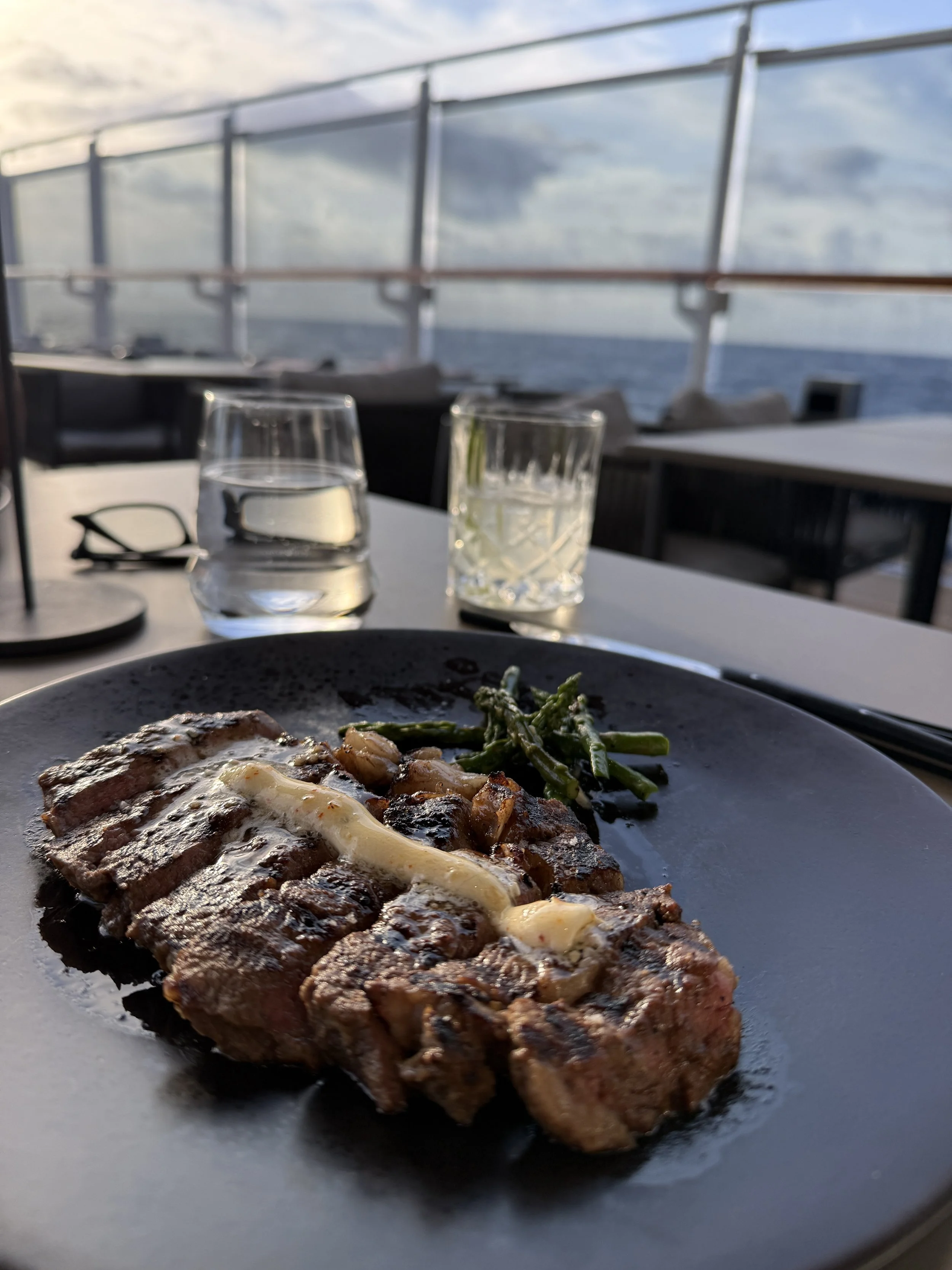A grilled steak topped with melting butter and a side of asparagus, served at an outdoor table aboard EXPLORA I with the ocean and sky behind it