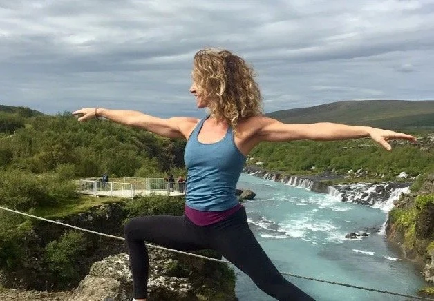 Yoga instructor Donna Zinna of Yoga World Traveler in Warrior II pose above a turquoise waterfall, arms outstretched