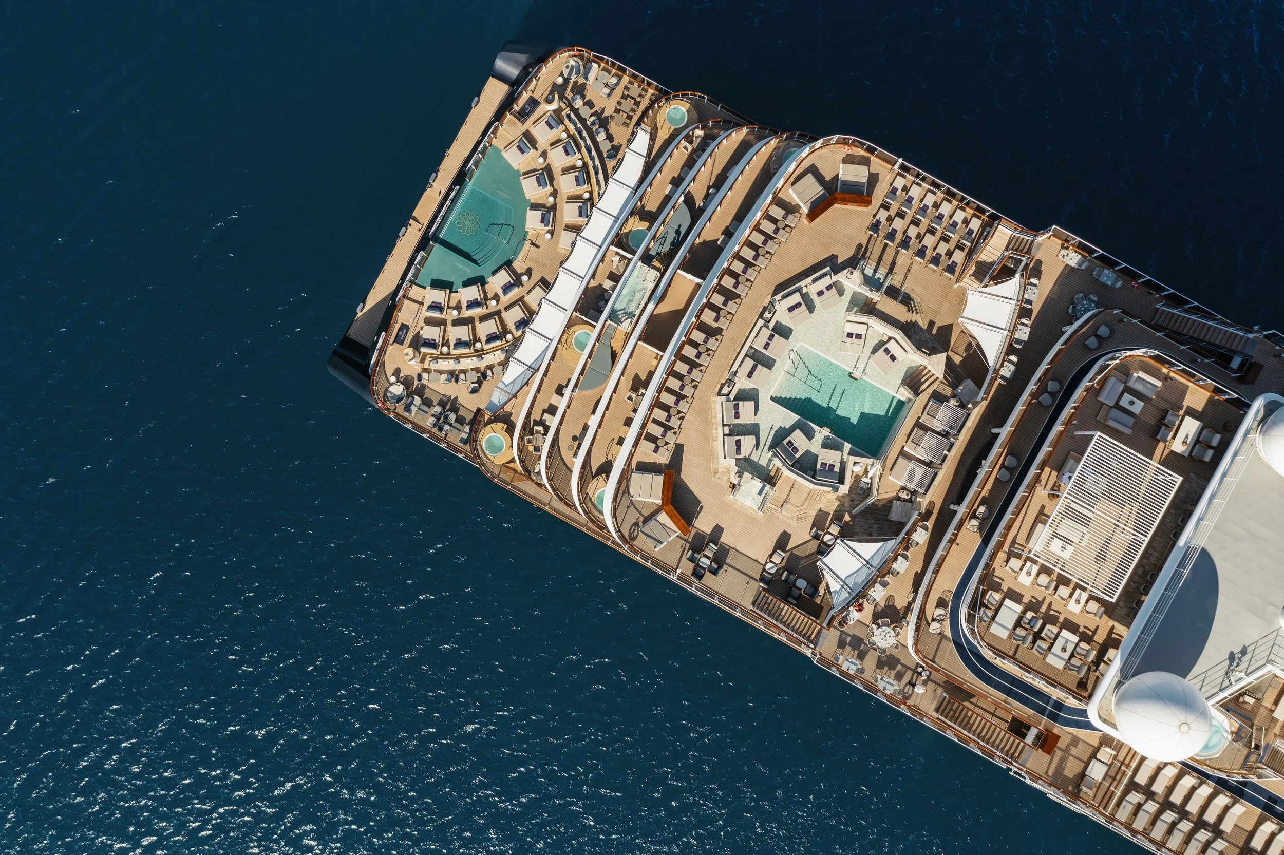 An overhead drone view of EXPLORA I at sea, showing the ship's layered pool decks, whirlpools, and rows of sun loungers surrounded by deep blue water