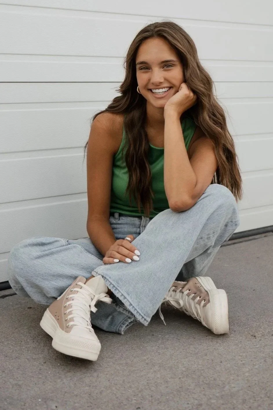 A young woman with long, wavy brown hair, smiling and sitting on the ground in front of a white wall. She is wearing a green sleeveless top, light blue oversized jeans, and beige high-top sneakers.