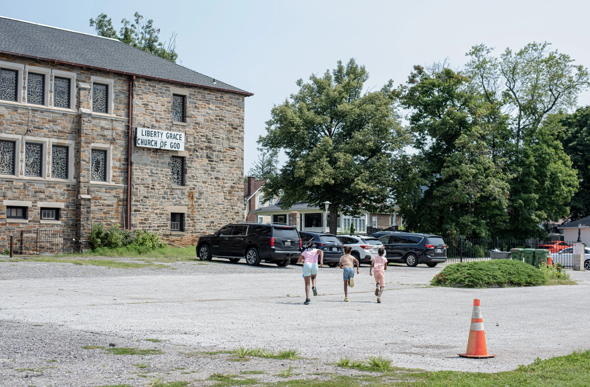 Three children running towards a church parking lot with cars parked and a large tree, in front of a stone church building with a sign that reads 'Liberty Grace Church of God'.