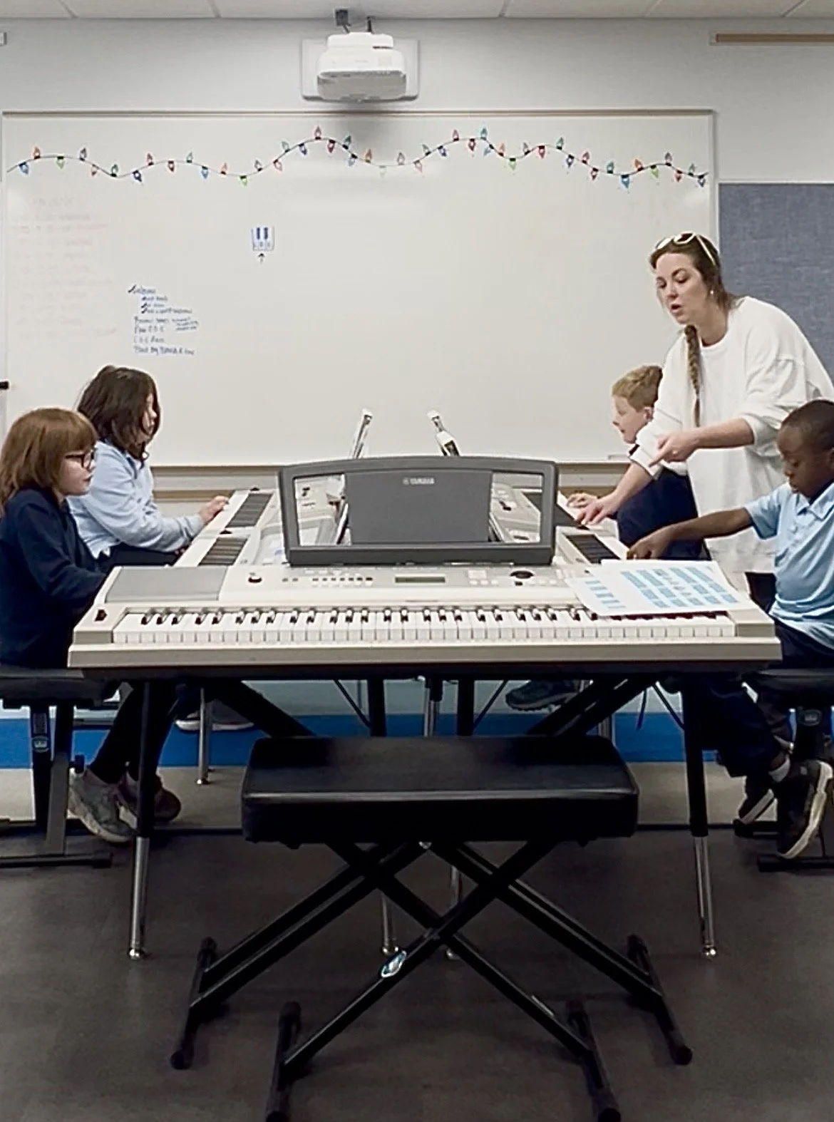 Children participating in a group piano class with an instructor at an electronic keyboard in a music classroom.
