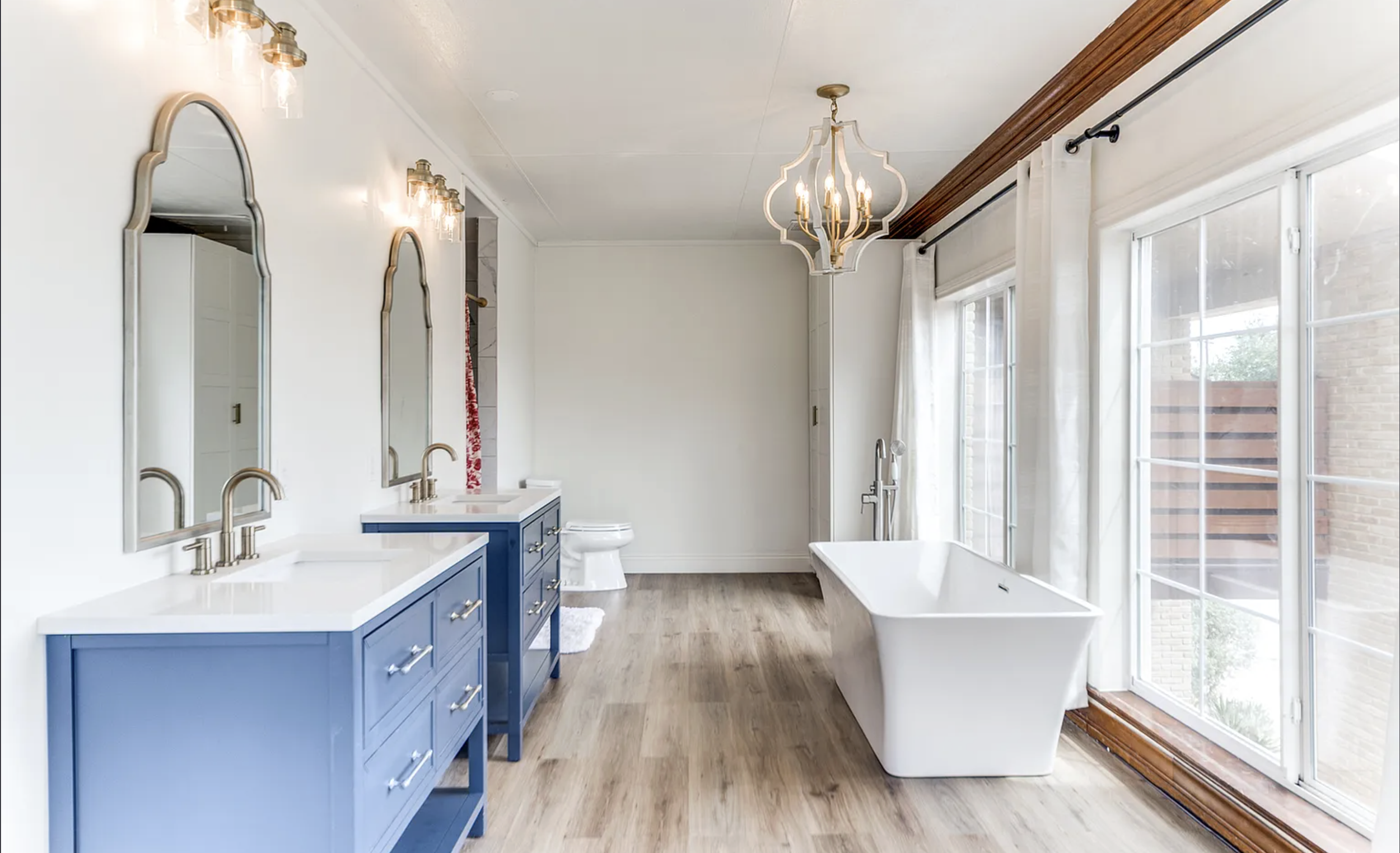 Bright bathroom with two blue vanities, each with a mirror and gold faucet, a white bathtub near large windows, and a modern chandelier hanging from the ceiling.