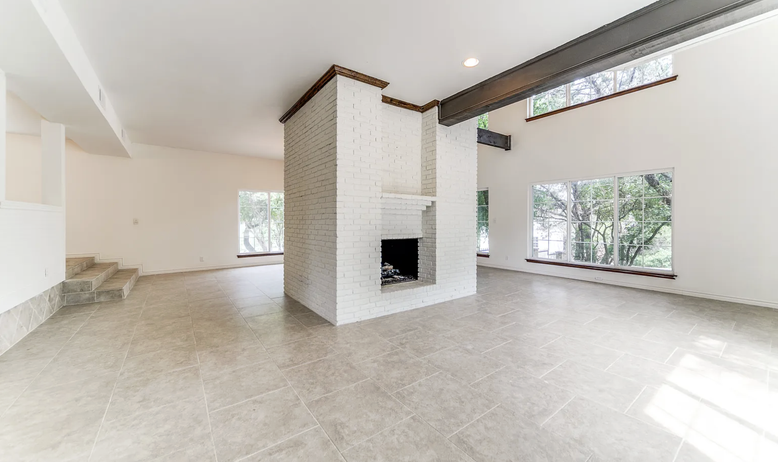 Empty living room with a white brick fireplace, large windows, tiled floor, and exposed ceiling beam.