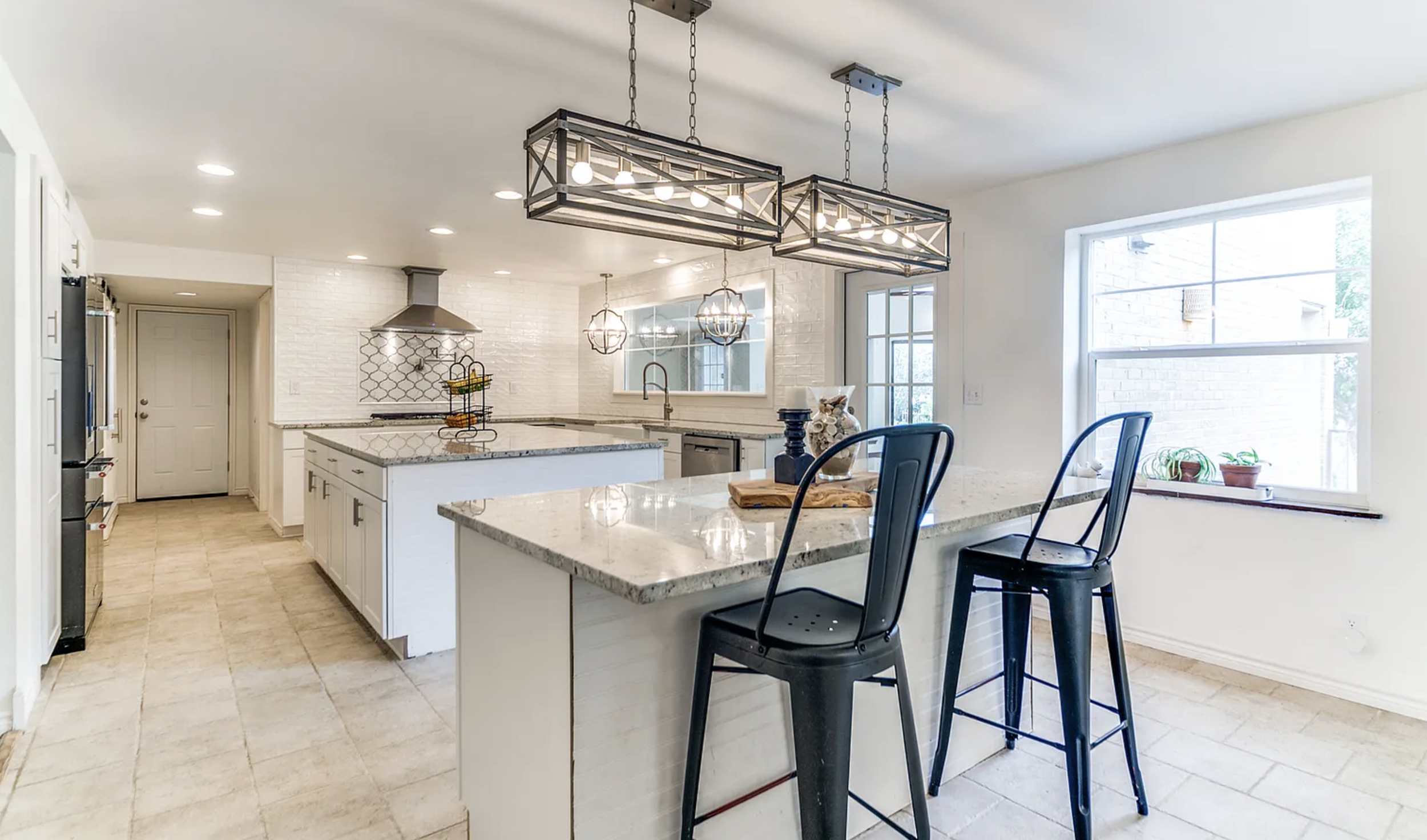 Bright kitchen with white cabinets, granite countertops, beige tile flooring, black barstools, and modern light fixtures, featuring a large window and stainless steel appliances.