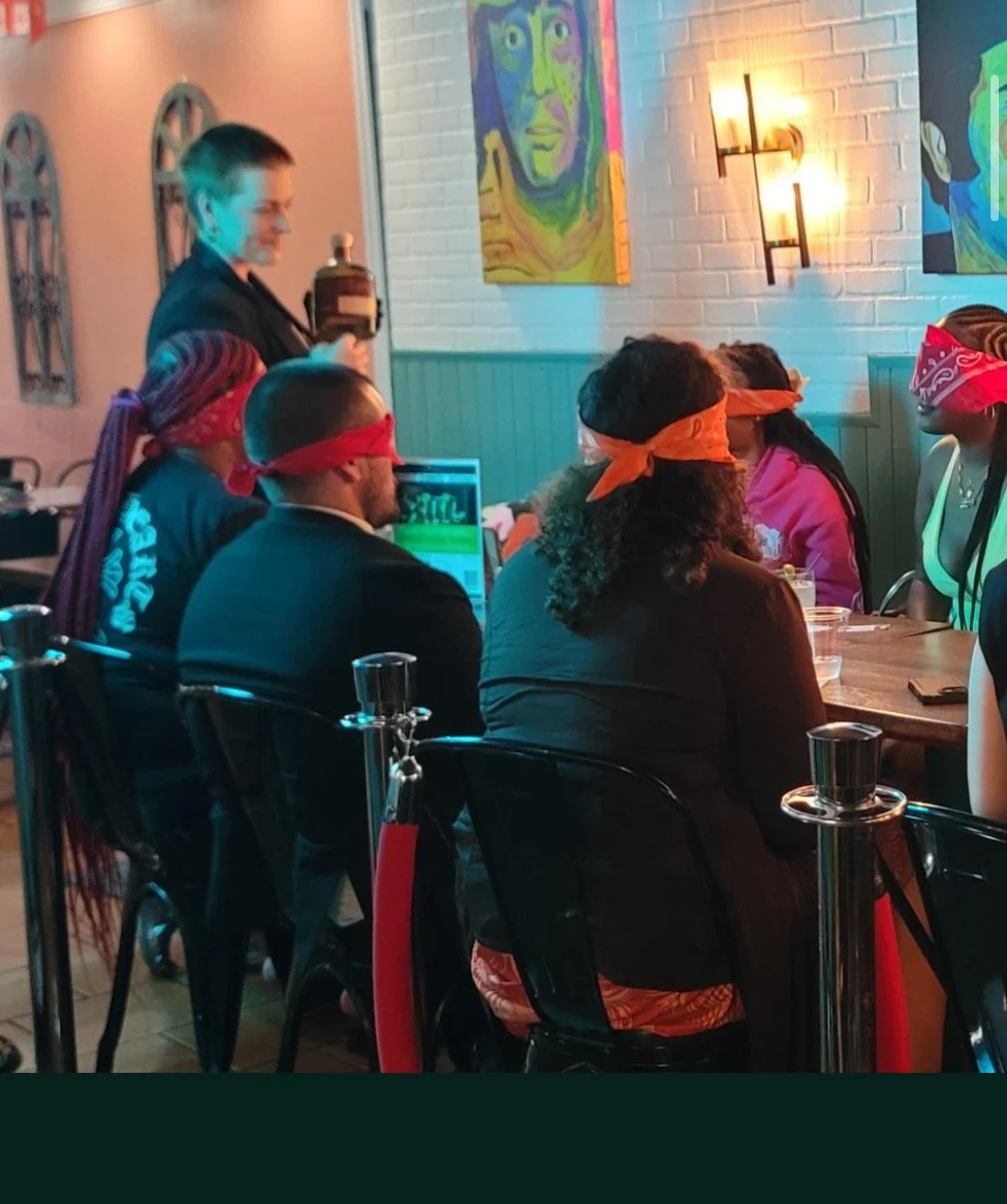 People participating in a themed event with their eyes covered by red and orange bandanas, seated at a table in a restaurant or bar. A person in a black shirt is standing and pouring a drink, with animated portraits on the white brick wall behind them.
