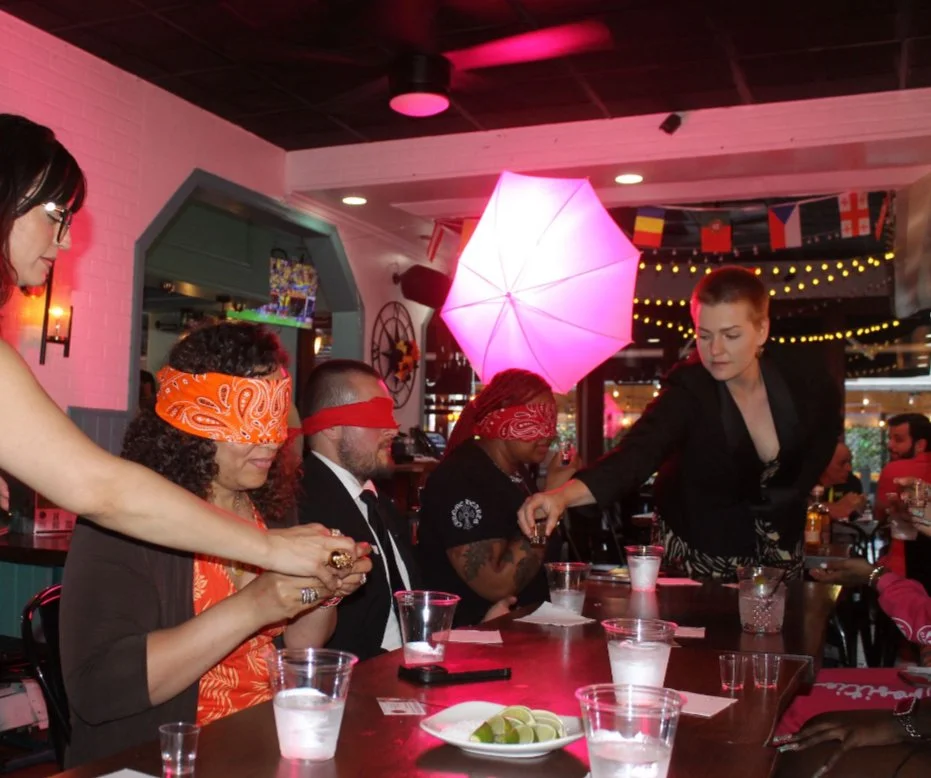 People at a bar participating in a blindfolded lime tasting game, with woman serving drinks and various flags and decorations in the background.
