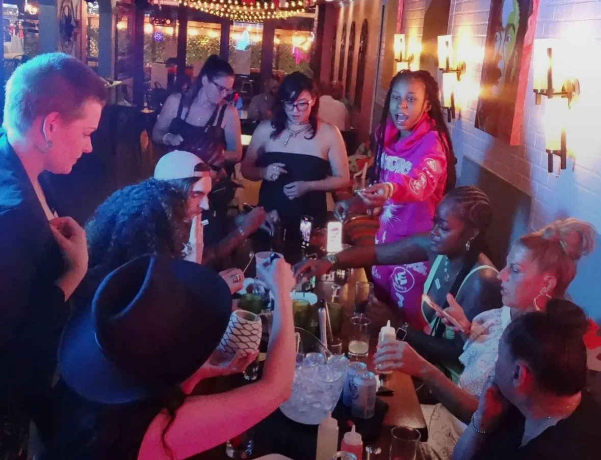 Group of people socializing at a lively indoor gathering, some seated at a table with drinks and snacks, others standing, illuminated by warm lighting and wall sconces, with colorful decorations and artwork on the walls.