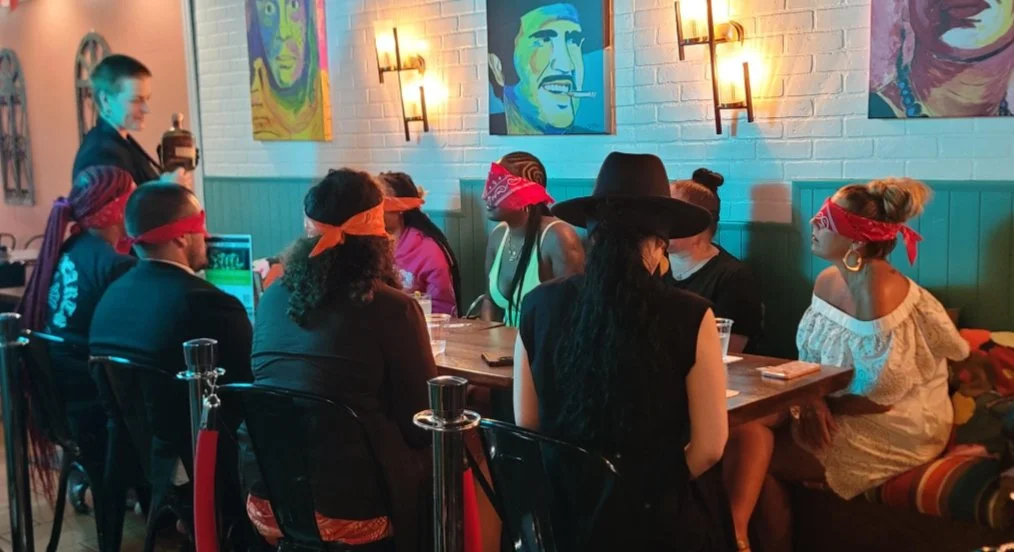 People sitting around a table at a restaurant, some wearing bandanas and one wearing a cowboy hat, with colorful paintings on the wall behind them.