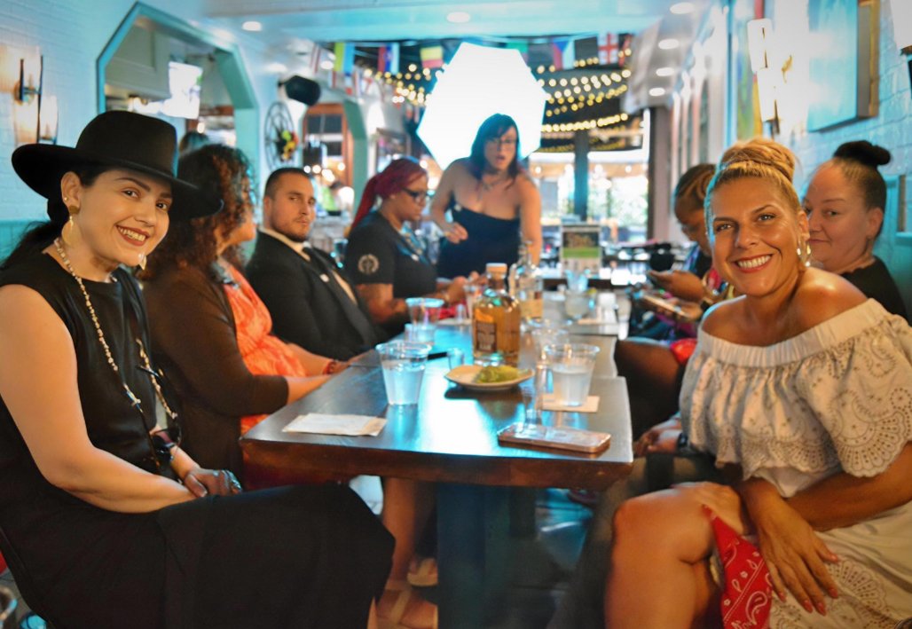 Group of people sitting at a long dining table inside a lively restaurant or bar, smiling at the camera, with colorful hanging decorations and string lights overhead.