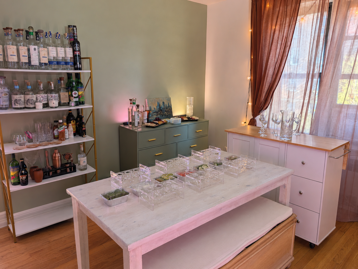 A dining room with a white wooden table holding empty clear acrylic containers, a green sideboard with bottles of alcohol, and a white cabinet with glasses on top. A tall shelf with liquor bottles stands beside the sideboard. The room has hardwood floors, green and white walls, sheer pink curtains, and string lights.