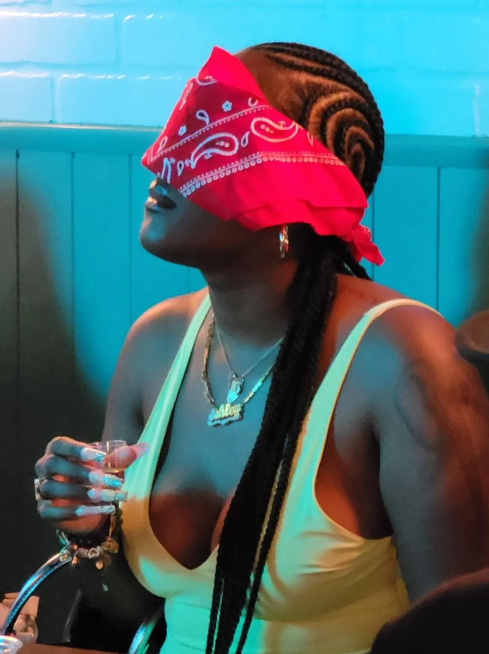 A woman with braided hair wearing a red bandanna over her eyes, a yellow tank top, layered necklaces, and multiple rings on her fingers, holding a glass in a dimly lit setting with blue brick walls in the background.