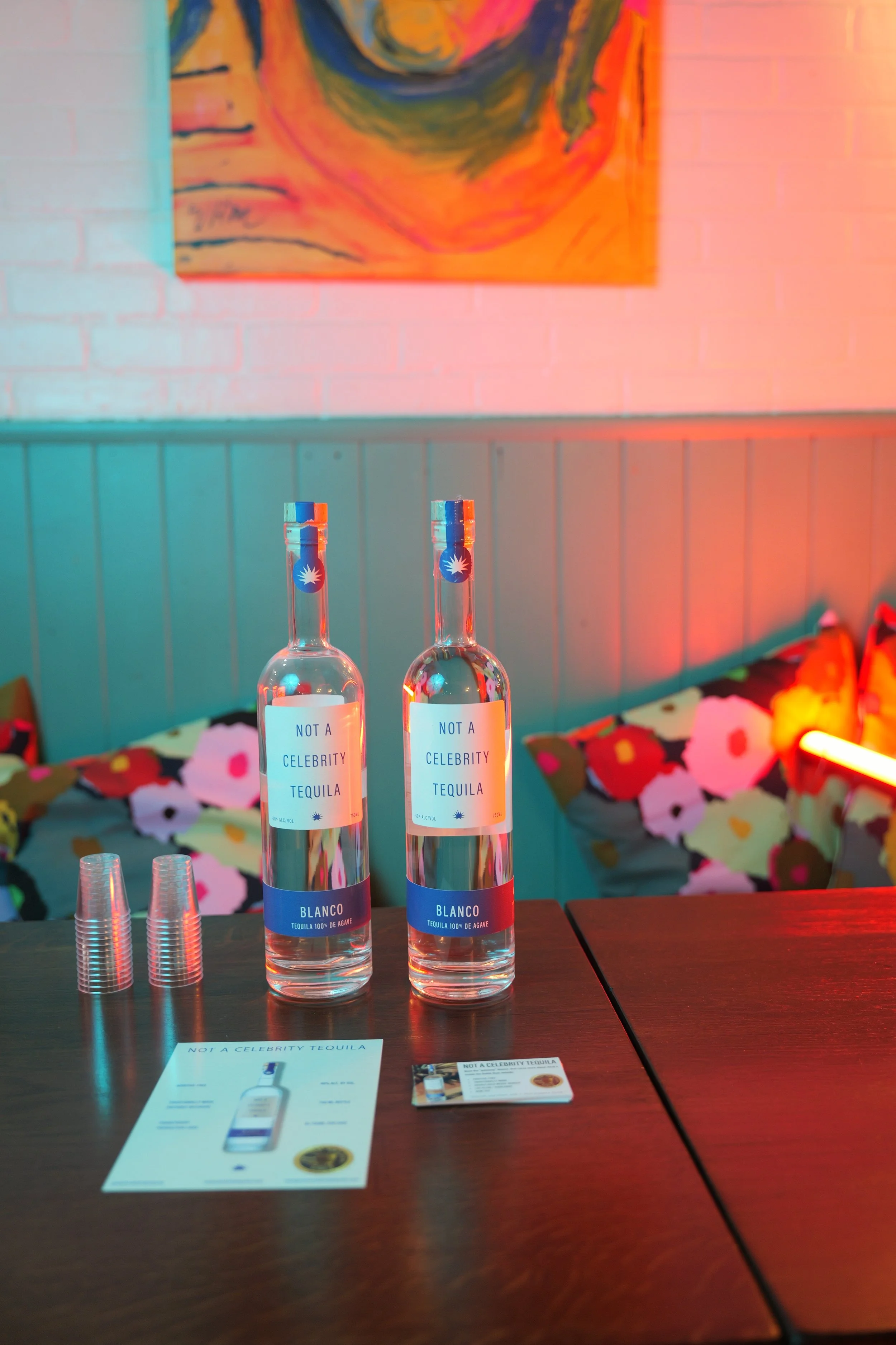 Two bottles of clear tequila labeled 'Not a Celebrity Tequila' with blue and red caps, placed on a dark wooden table. Behind them, there are clear plastic cups, promotional flyers, and a blurred background with a colorful abstract painting and some flower arrangements.