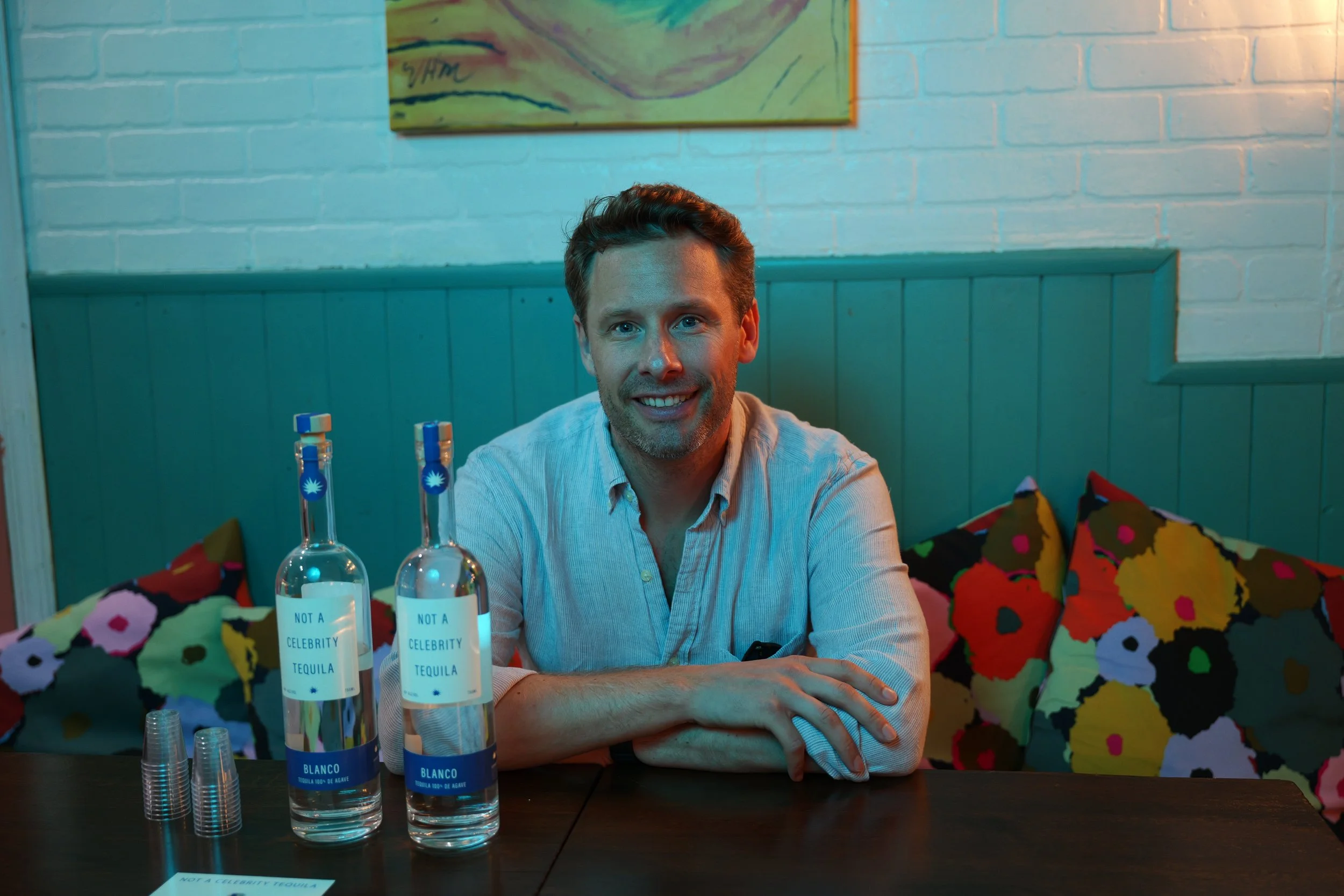 A man with light skin, brown hair, and a light beard, smiling at the camera, sitting at a table inside a colorful restaurant. In front of him, there are three bottles of tequila with labels reading "NOT A CELEBRITY TEQUILA" and "BLANCO," and a stack of clear plastic shot glasses.